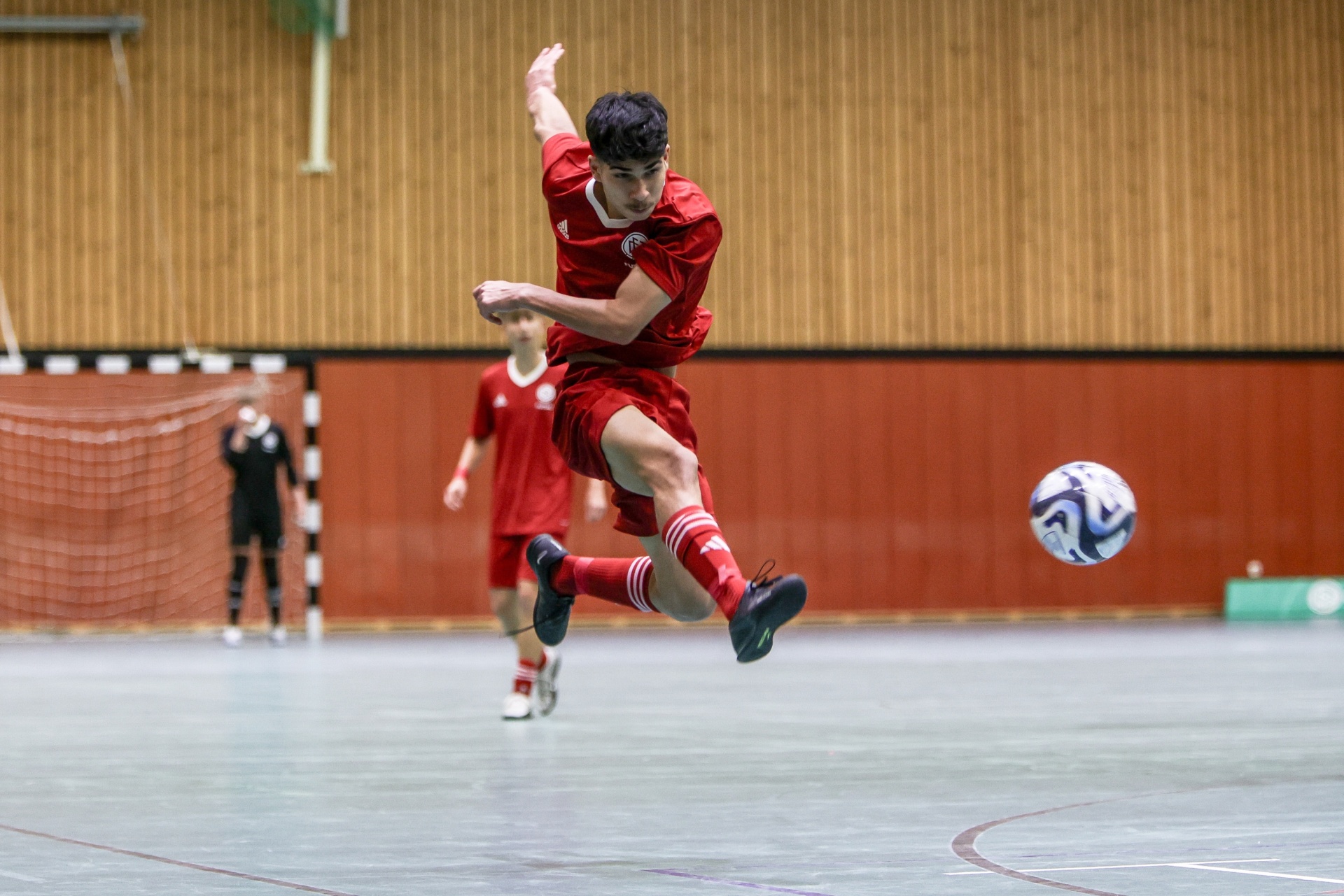 [{"clang_id":1,"value":"Futsalspieler in Rot springt beim Schießen auf Ball zu. Im Hintergrund zwei weitere Spieler und ein Tor. Hallenfußballfeld mit Holzwand."}]