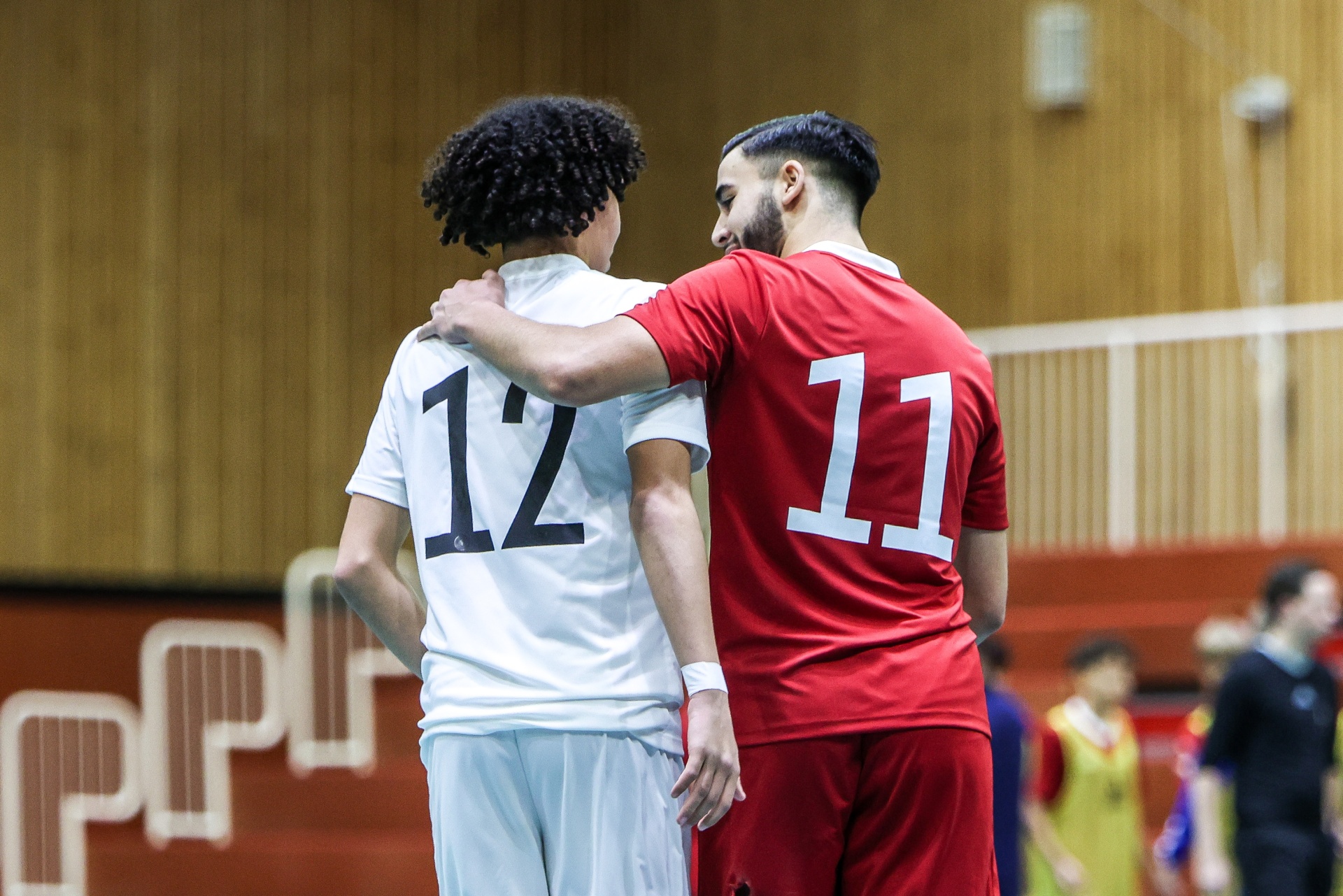 [{"clang_id":1,"value":"Zwei Futsalspieler mit den Rückennummern 11 und 12 stehen Rücken an Rücken in einer Sporthalle. Im Hintergrund verschwommene Zuschauer."}]