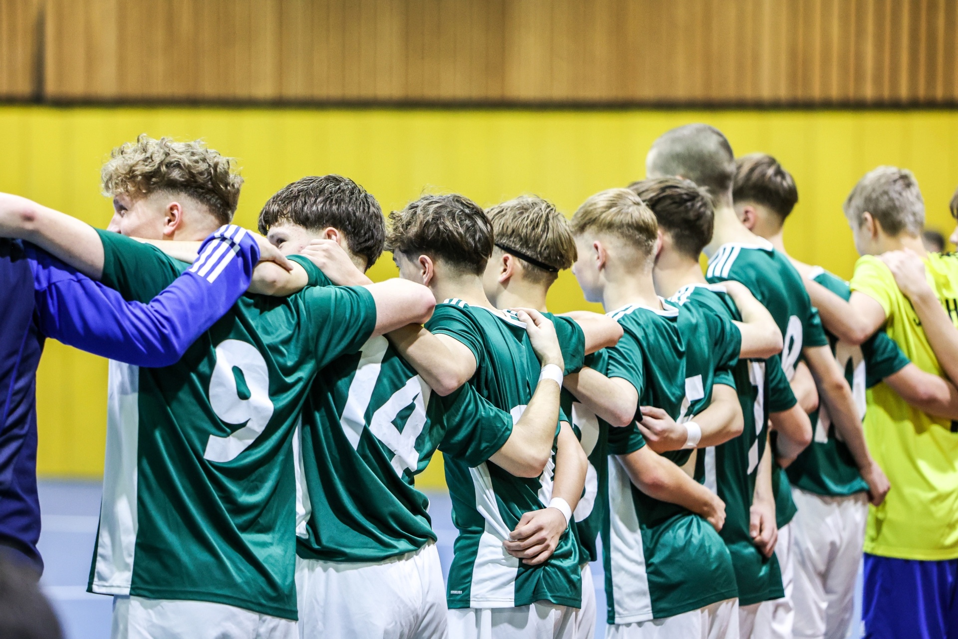 [{"clang_id":1,"value":"Fussballmannschaft in grünen Trikots mit Arm um die Schulter, Reihe stehend, Blick nach unten. Im Hintergrund gelbe Wand und gegnerische Spieler in gelben Trikots."}]