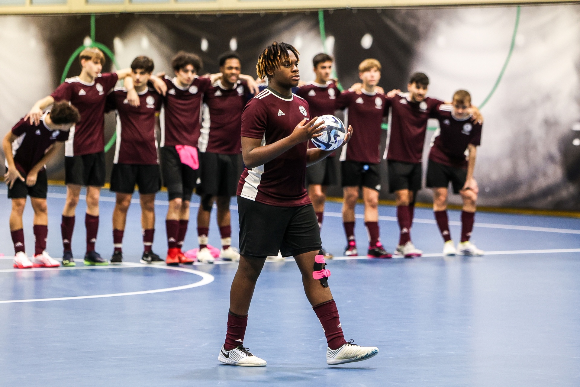 [{"clang_id":1,"value":"Futsalteam: Ein einzelner Spieler mit Ball im Vordergrund, dahinter eine Reihe von Teammitgliedern in Vereinskleidung in einer Sporthalle."}]