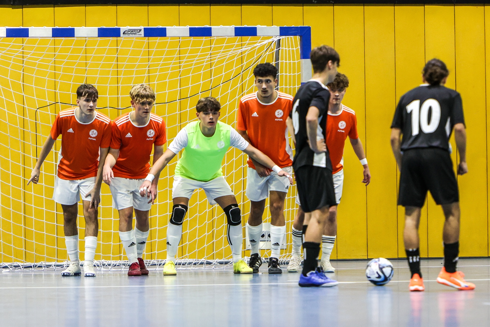 [{"clang_id":1,"value":"Futsal-Spiel: Torwart in grüner Trainingsweste, fünf Spieler in roten Trikots und ein Spieler mit Nummer 10 in schwarzen Shorts stehen auf blauem Hallenboden vor einem Tor."}]