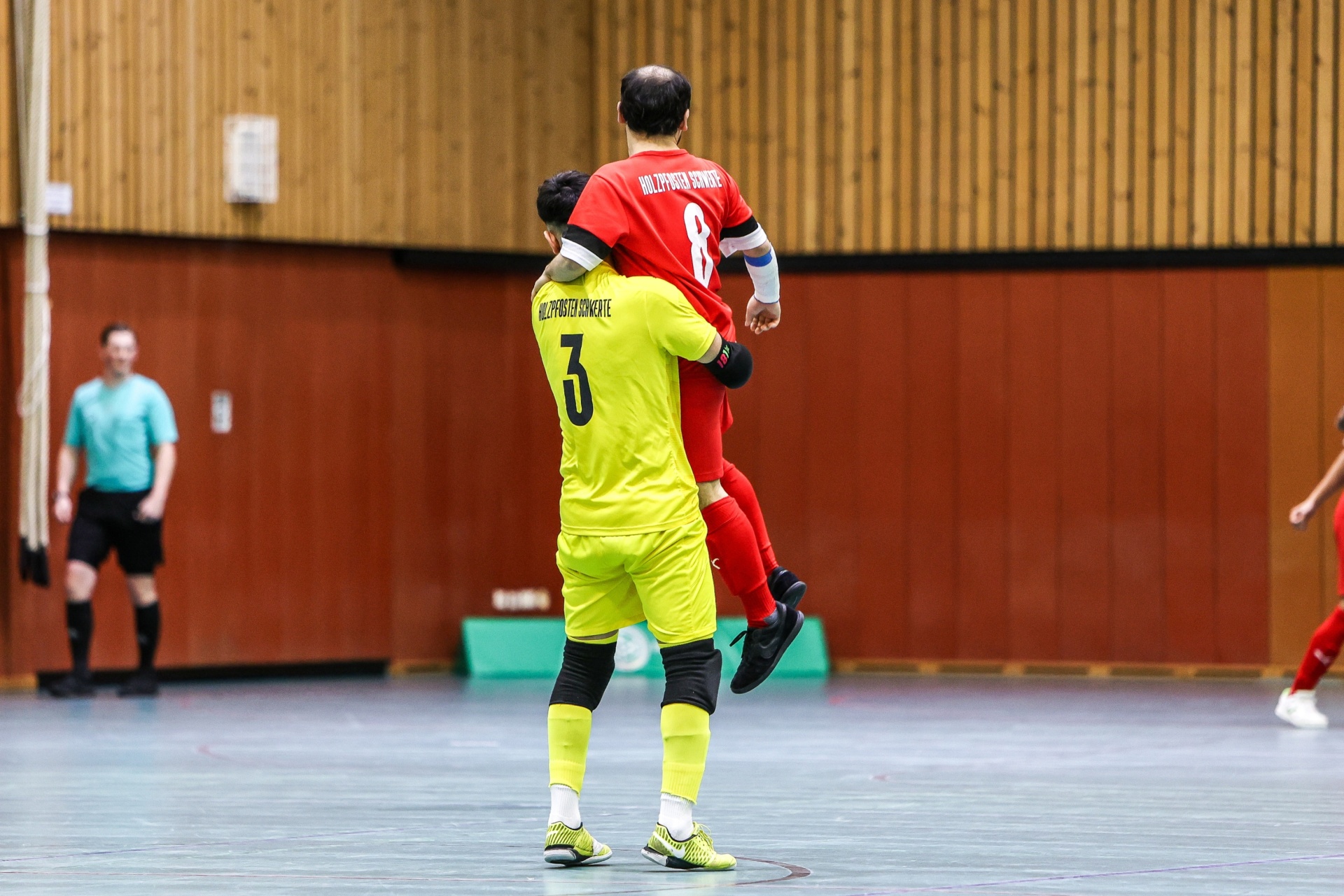 [{"clang_id":1,"value":"Zwei Futsalspieler feiern: Spieler mit Trikotnummer 3 trägt Spieler mit Trikotnummer 8 auf seinen Schultern. Im Hintergrund weitere Spieler und eine Holzwand."}]