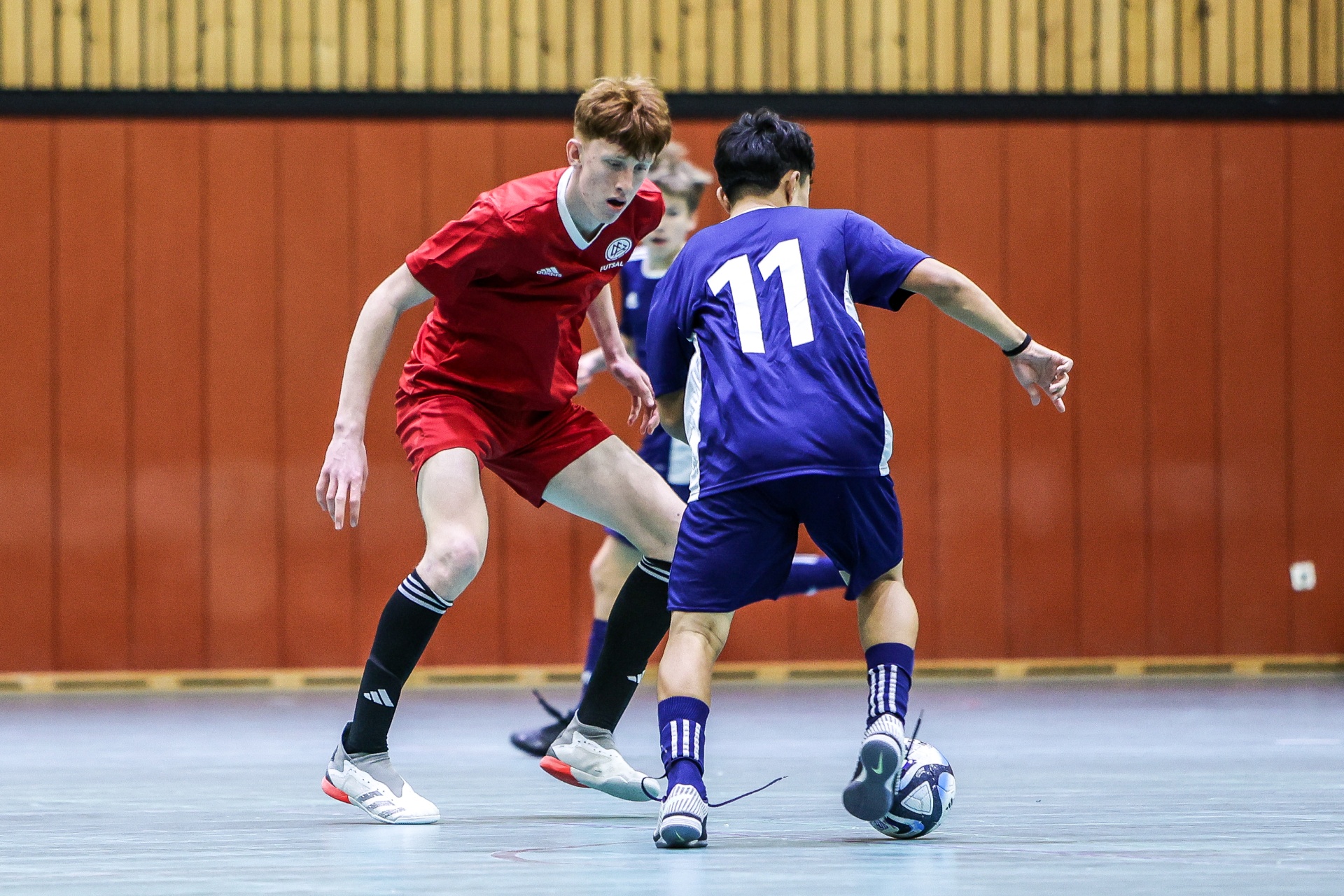[{"clang_id":1,"value":"Zwei Futsalspieler im Wettkampf. Links ein Spieler in rot, rechts in blau mit Nummer 11, beide konzentriert auf den Ball."}]