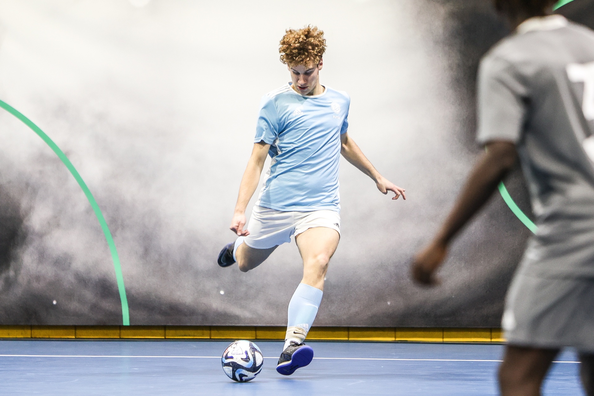 [{"clang_id":1,"value":"Junger Fußballspieler mit blonden Locken schießt im Hallenfußball auf das Tor. Gegner im Hintergrund. Blaue Sporthalle, grüner Rand."}]