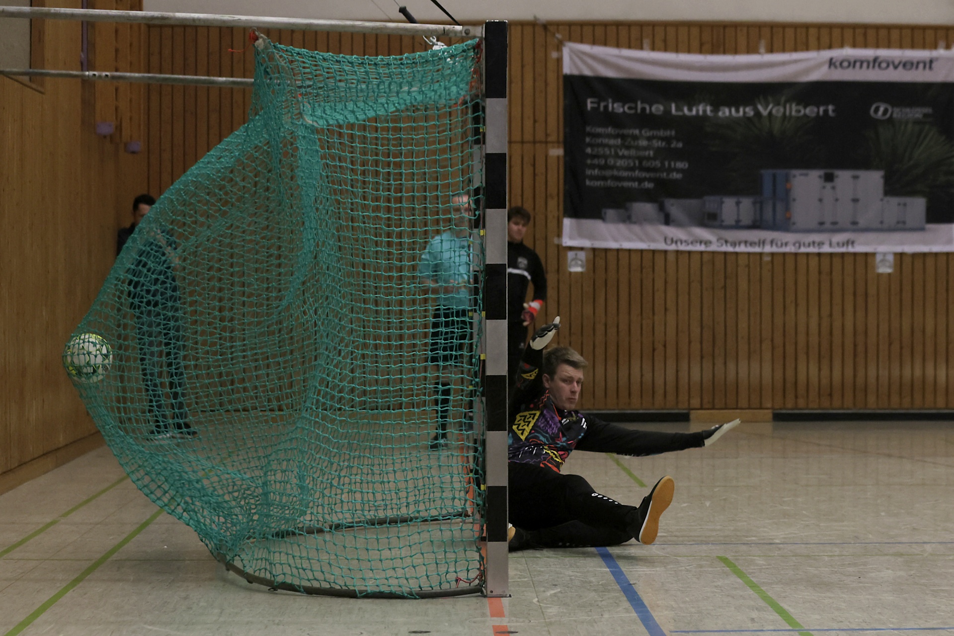 [{"clang_id":1,"value":"Handballtor mit Netz, davor liegender Torwart in Trainingsmontur, linke Hand ausgestreckt. Hintergrund: Sporthalle mit Werbebanner."}]