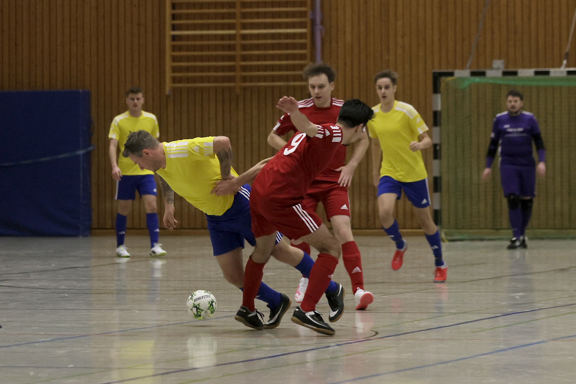 [{"clang_id":1,"value":"Futsalspiel in Sporthalle: Zwei Spieler kämpfen um den Ball. Im Hintergrund weitere Spieler in gelben und violetten Trikots."}]