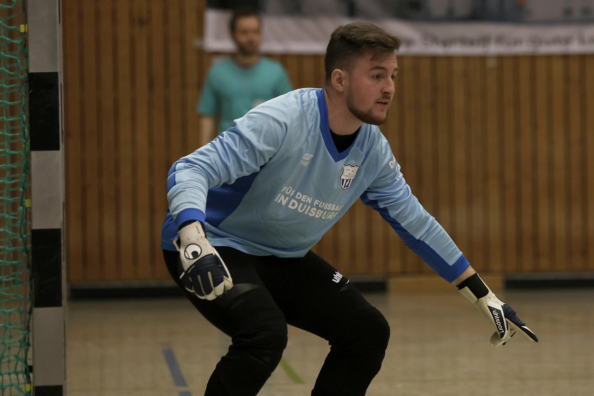 [{"clang_id":1,"value":"Fußballtorwart in hellblauer Ausrüstung kauert auf einem Hallenfußballfeld. Im Hintergrund eine Holzwand und eine verschwommene Person."}]