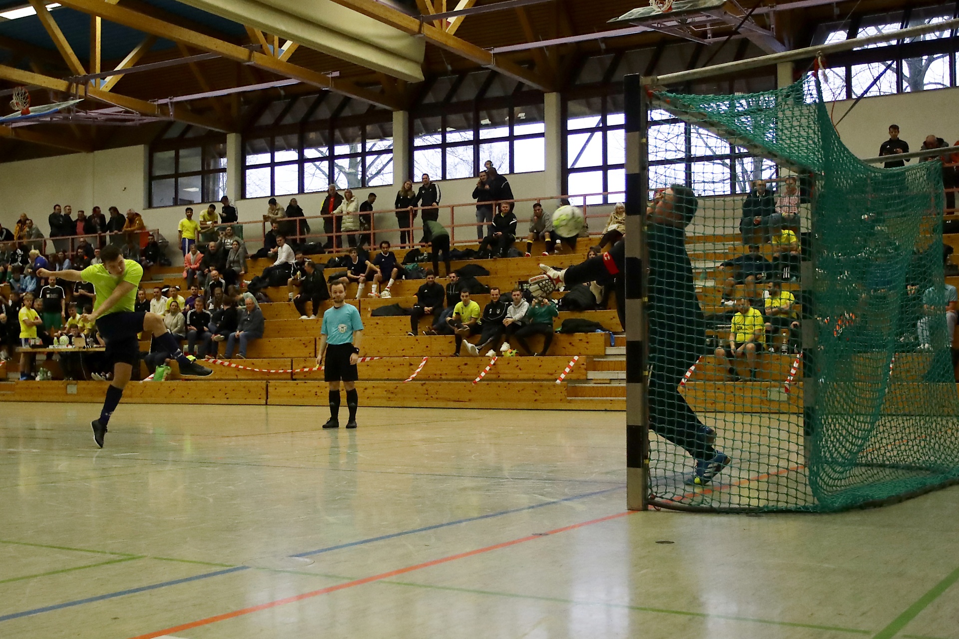 [{"clang_id":1,"value":"Handballspiel in Sporthalle. Spieler in gelben Trikots greifen an, Gegenspieler am Tor, Schiedsrichter dazwischen, Zuschauer auf Tribüne."}]