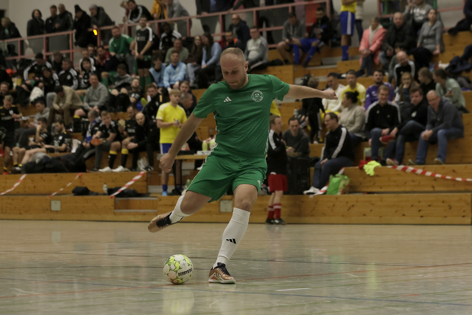 [{"clang_id":1,"value":"Futsalspieler in grüner Sportkleidung schießt Ball in einer Sporthalle. Im Hintergrund Zuschauer auf Tribünen."}]