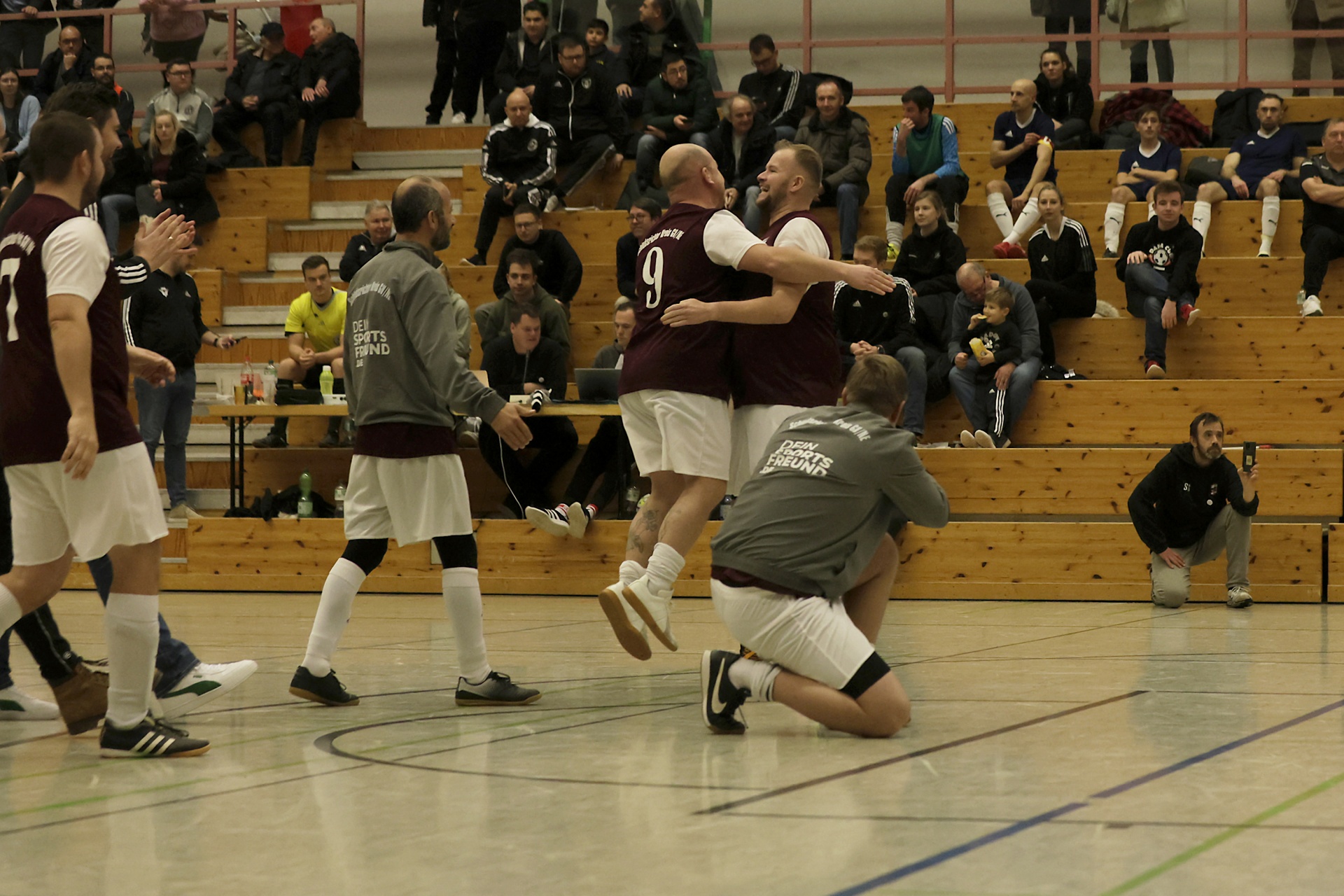 [{"clang_id":1,"value":"Handballspiel in Sporthalle. Zwei Spieler in Dunkelrot und Weiß feiern mit erhobenen Armen. Zuschauerreihen im Hintergrund."}]