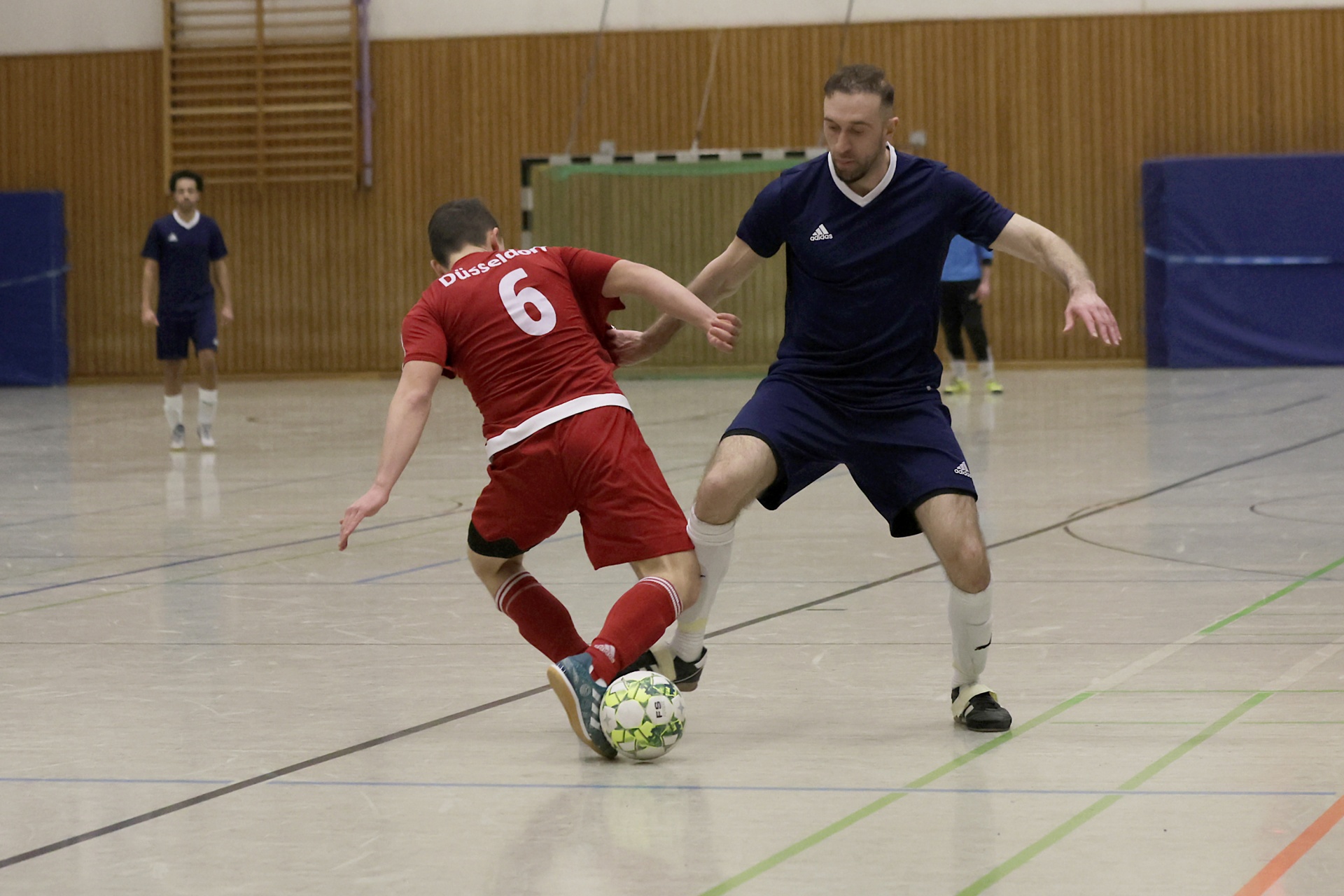 [{"clang_id":1,"value":"Zwei Futsalspieler im Hallensport: Ein Spieler in Rot dribbelt den Ball, wird von einem Spieler in Dunkelblau gedeckt. Im Hintergrund weitere Spieler und eine Sporthalle."}]