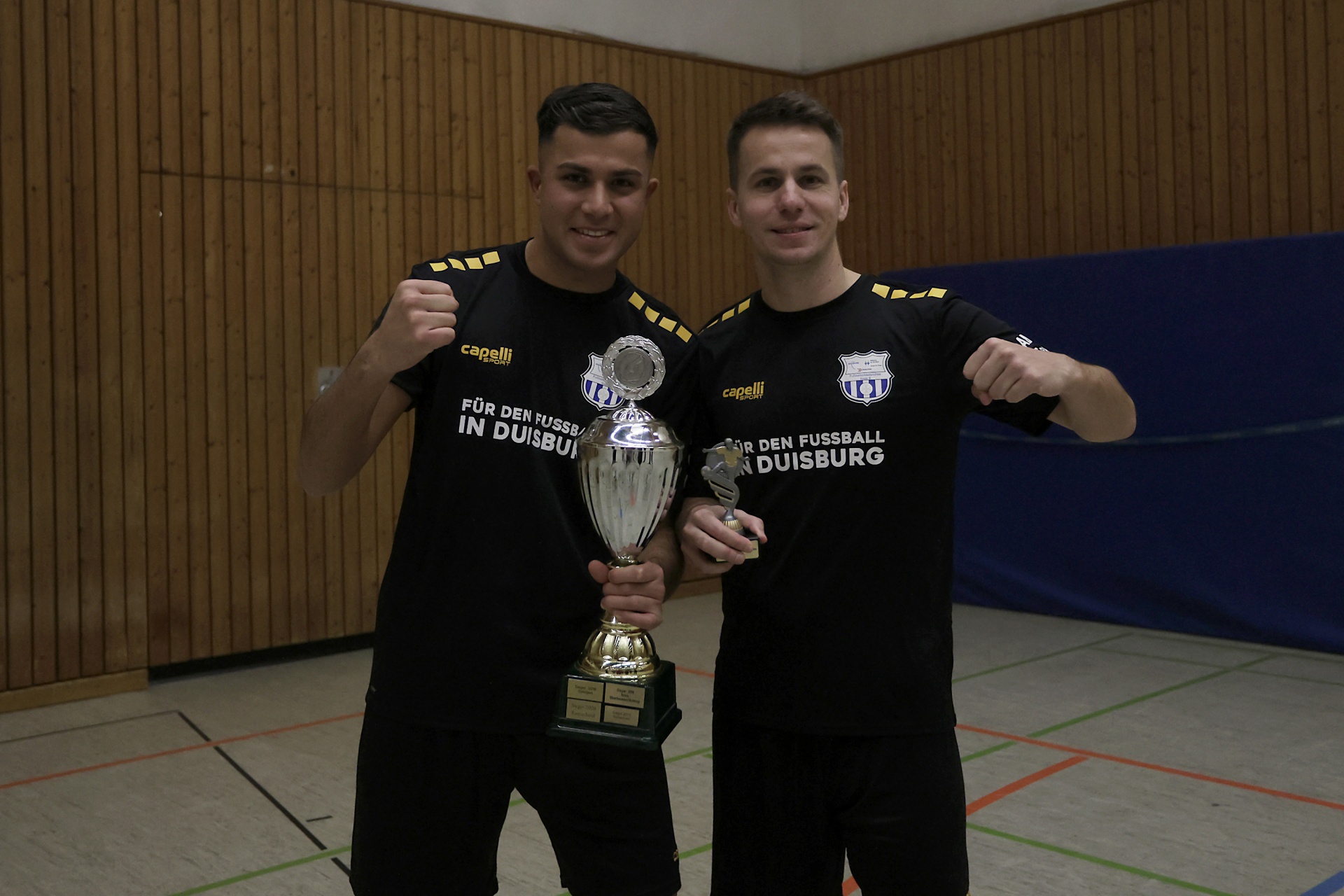 [{"clang_id":1,"value":"Zwei Männer in schwarzen Sportshirts mit Aufdruck halten Pokale in einer Sporthalle. Beide blicken in die Kamera und zeigen ihre Muskeln."}]
