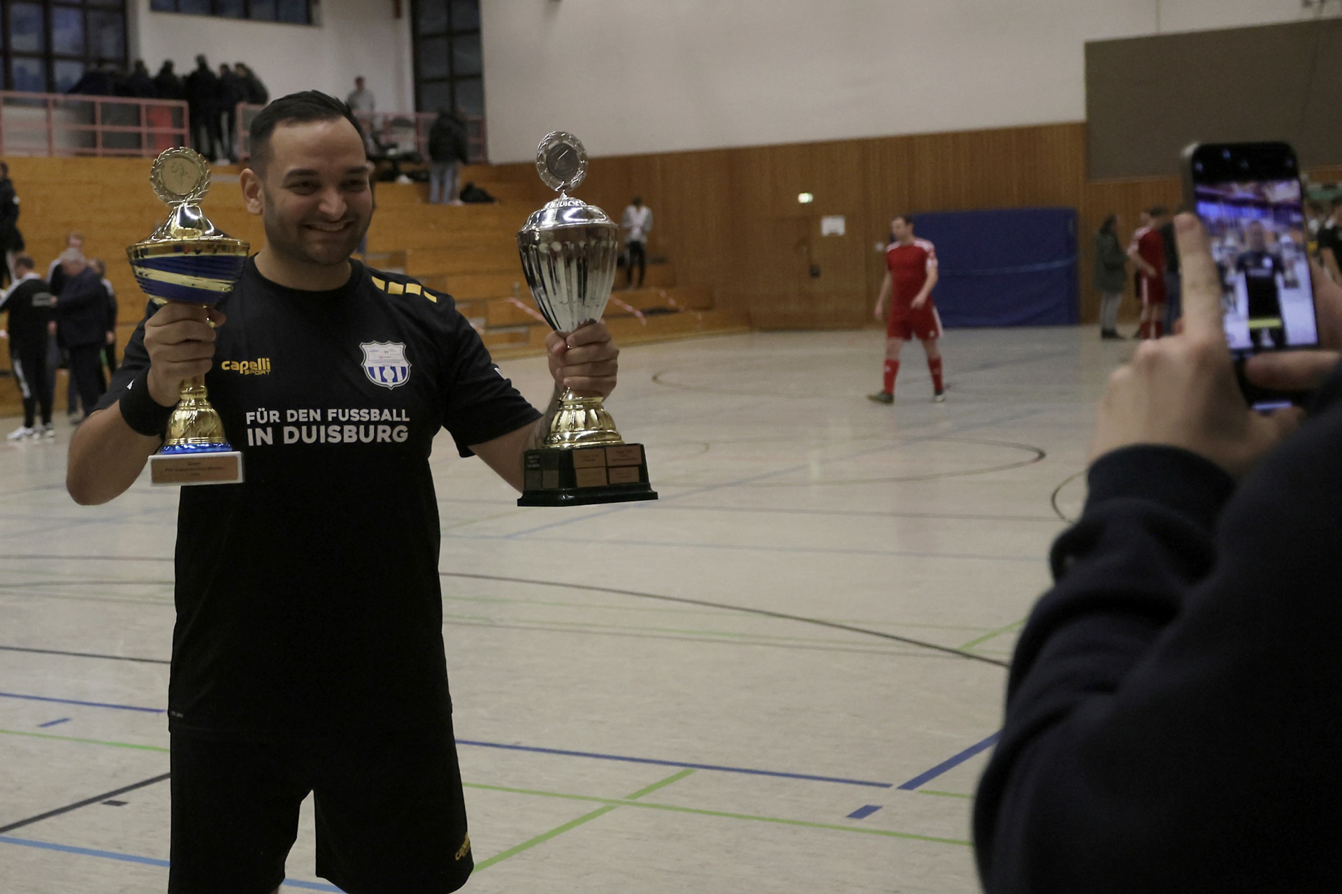 [{"clang_id":1,"value":"Mann mit zwei Trophäen in einer Sporthalle. Im Hintergrund Personen und ein Mann in roten Sportkleidern. Jemand filmt mit einem Smartphone."}]
