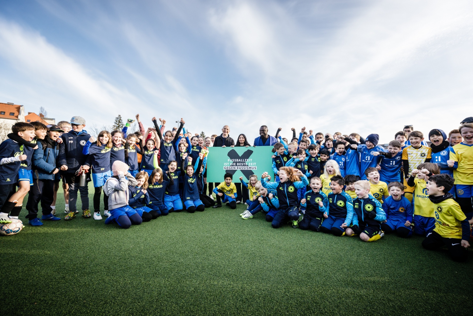 [{"clang_id":1,"value":"Fußballmannschaft mit Trainer posiert auf grünem Kunstrasenfeld. Kinder und Jugendliche tragen blaue und gelbe Trikots, einige heben die Arme. Im Hintergrund Gebäude und bewölkter Himmel."}]
