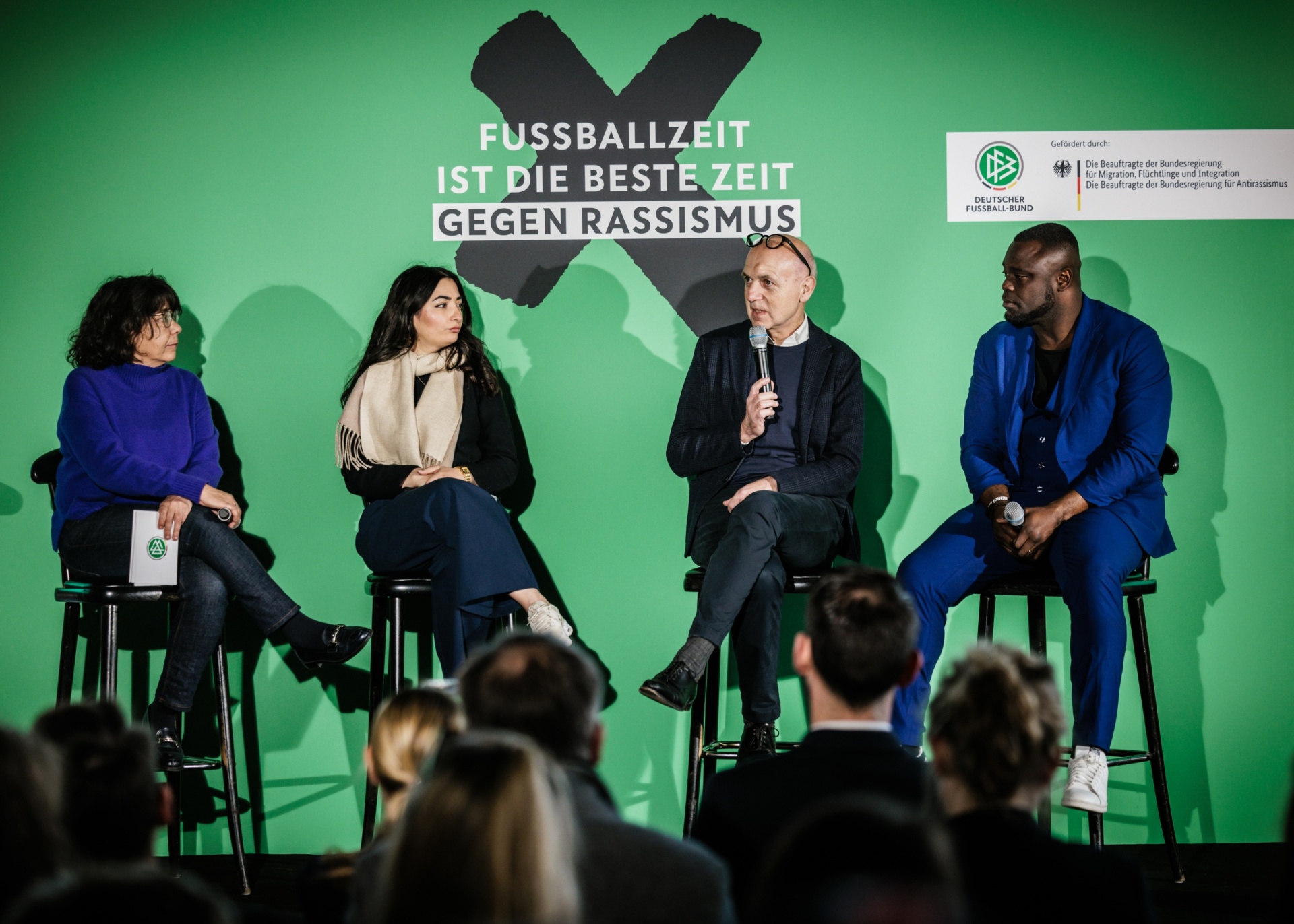 [{"clang_id":1,"value":"Paneldiskussion: Vier Personen sitzen auf Stühlen vor grünem Hintergrund mit Aufschrift gegen Rassismus. Publikum im Vordergrund."}]