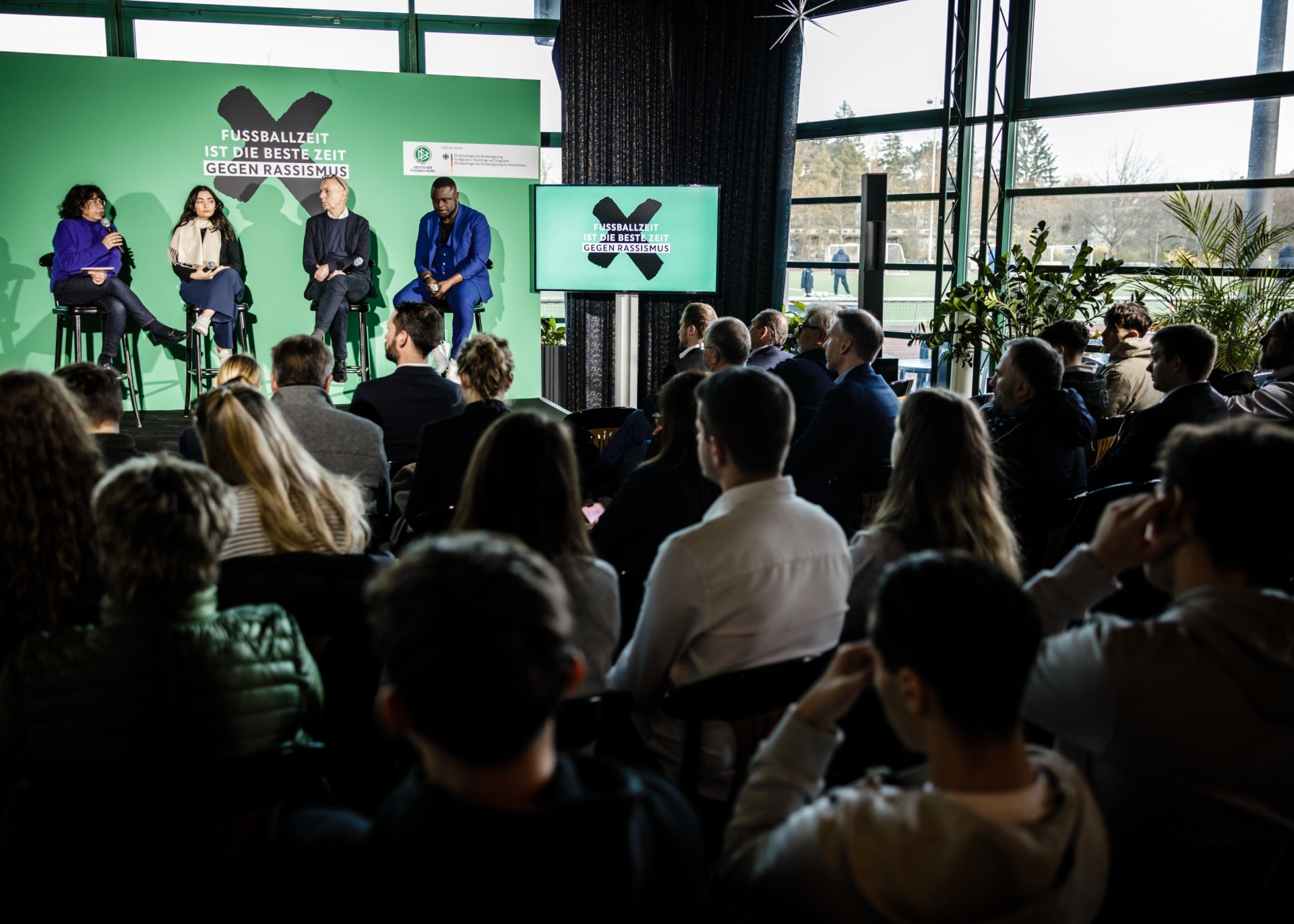 [{"clang_id":1,"value":"Paneldiskussion mit fünf Personen am Tisch, vor Publikum. Im Hintergrund ein Banner mit Aufschrift \"Fussballzeit ist die beste Zeit gegen Rassismus\". Großer Raum mit Fenstern."}]