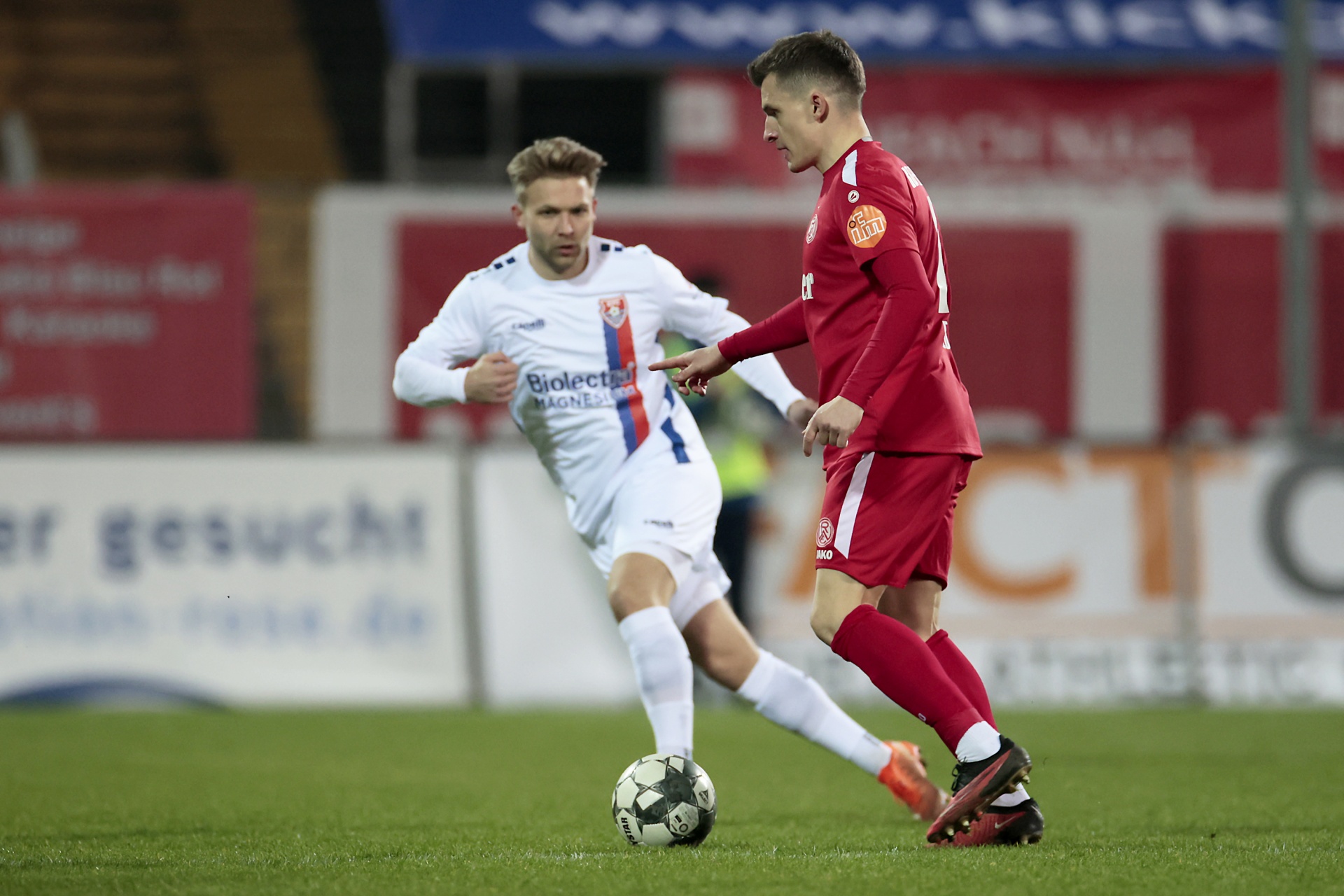 [{"clang_id":1,"value":"Zwei Fußballspieler auf dem Rasen. Ein Spieler in Rot dribbelt den Ball, der andere in Weiß und Blau folgt ihm. Im Hintergrund ein Stadion."}]