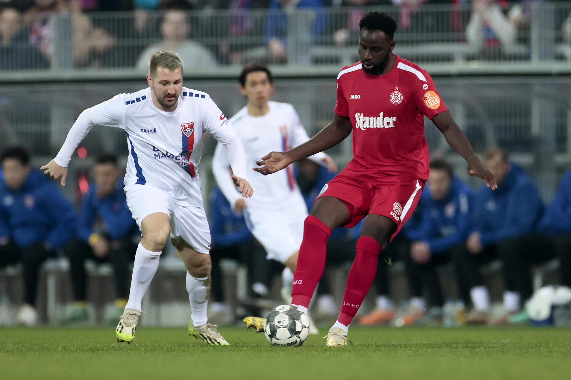 [{"clang_id":1,"value":"Zwei Fußballspieler im Spiel. Links ein Spieler mit weißem Trikot, rechts ein Spieler mit rotem Trikot und Ball. Im Hintergrund Zuschauer."}]