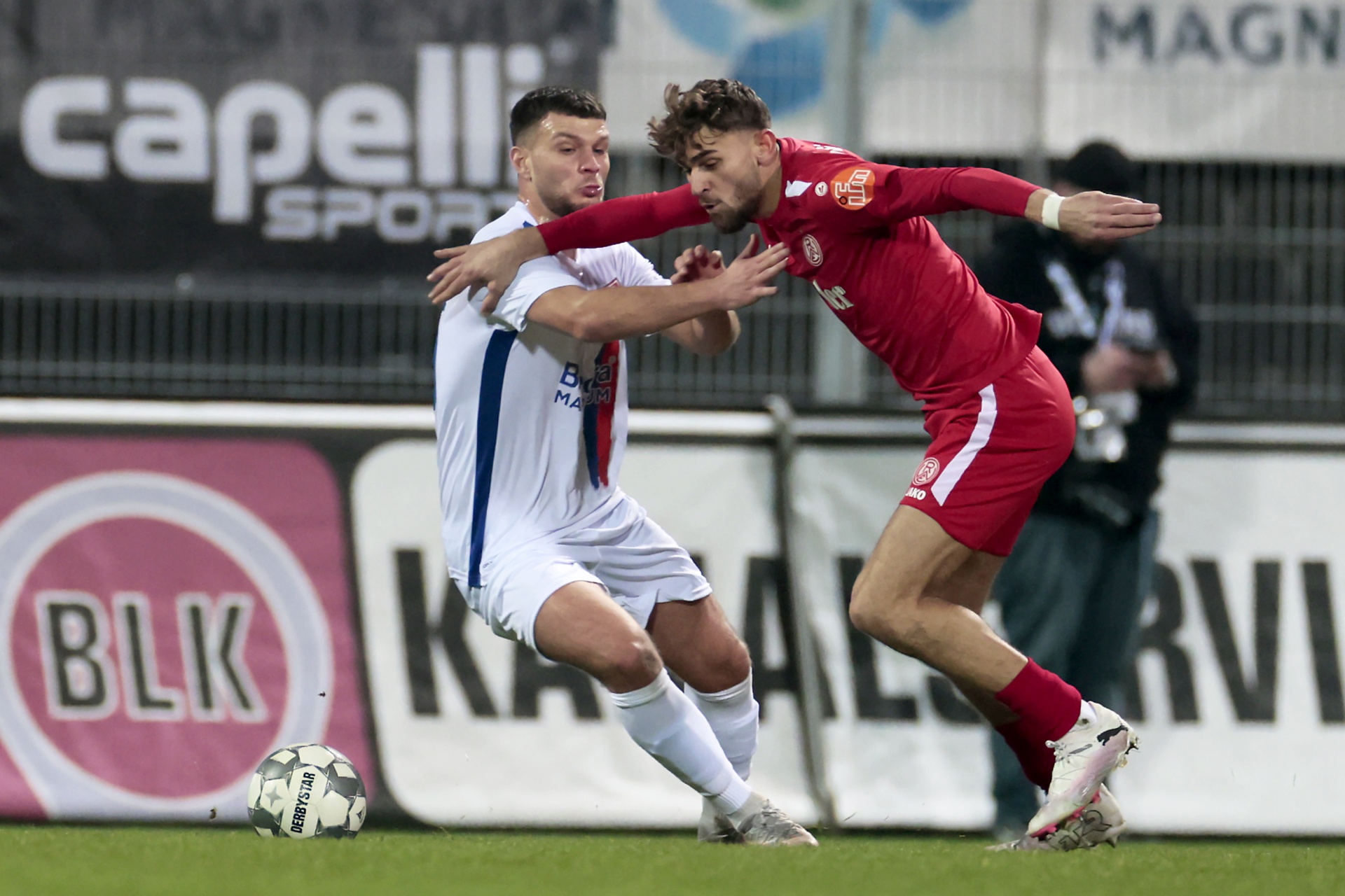 [{"clang_id":1,"value":"Zwei Fußballspieler kämpfen um den Ball auf dem Rasen. Links ein Spieler in blau-weißem Trikot, rechts in rotem Trikot. Im Hintergrund Werbebanner und Zuschauer."}]