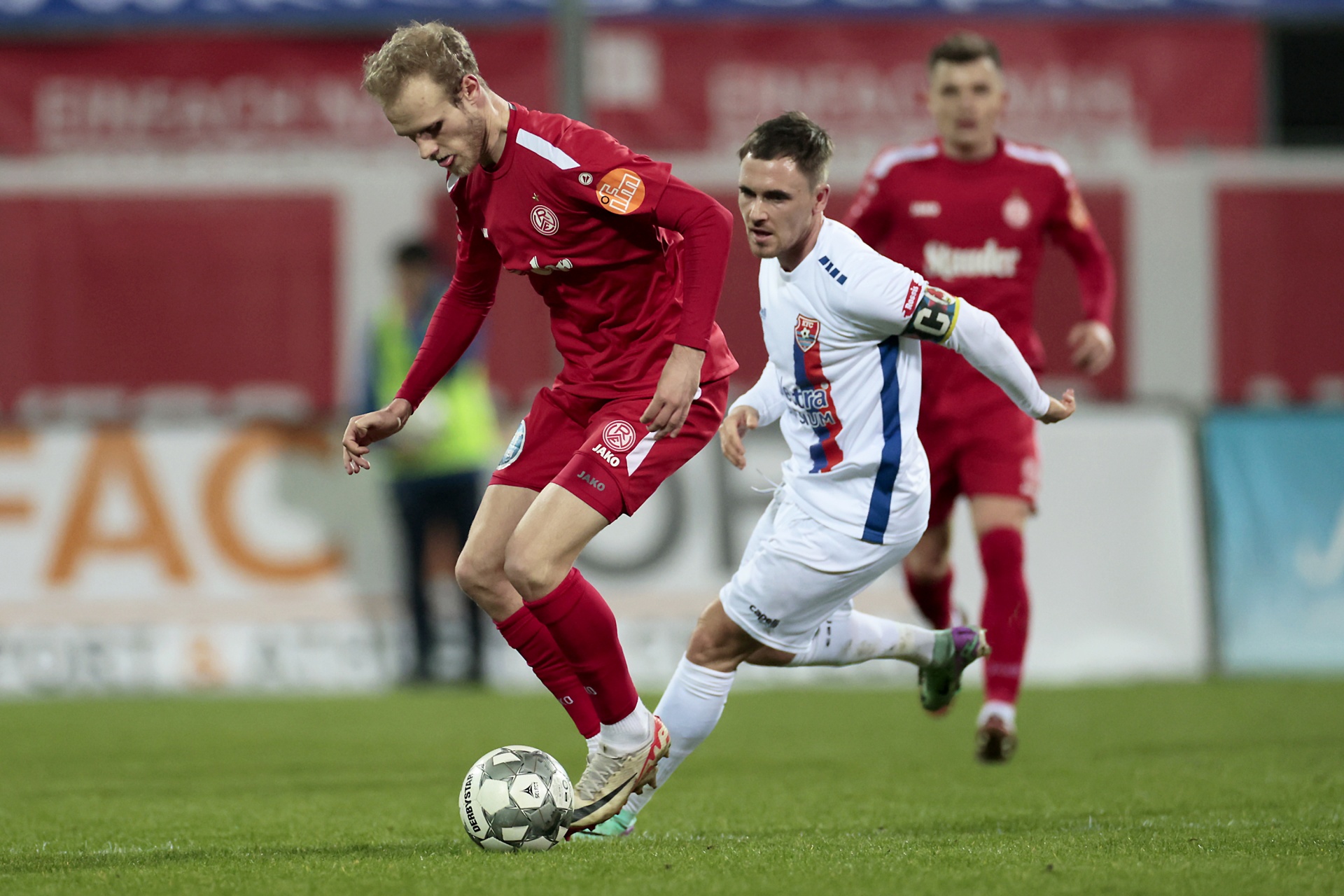 [{"clang_id":1,"value":"Zwei Fußballspieler im Spiel. Links ein Spieler in roten Trikots, der den Ball spielt. Rechts ein Spieler in weiß-blauem Trikot, der ihn verfolgt. Im Hintergrund weitere Spieler."}]