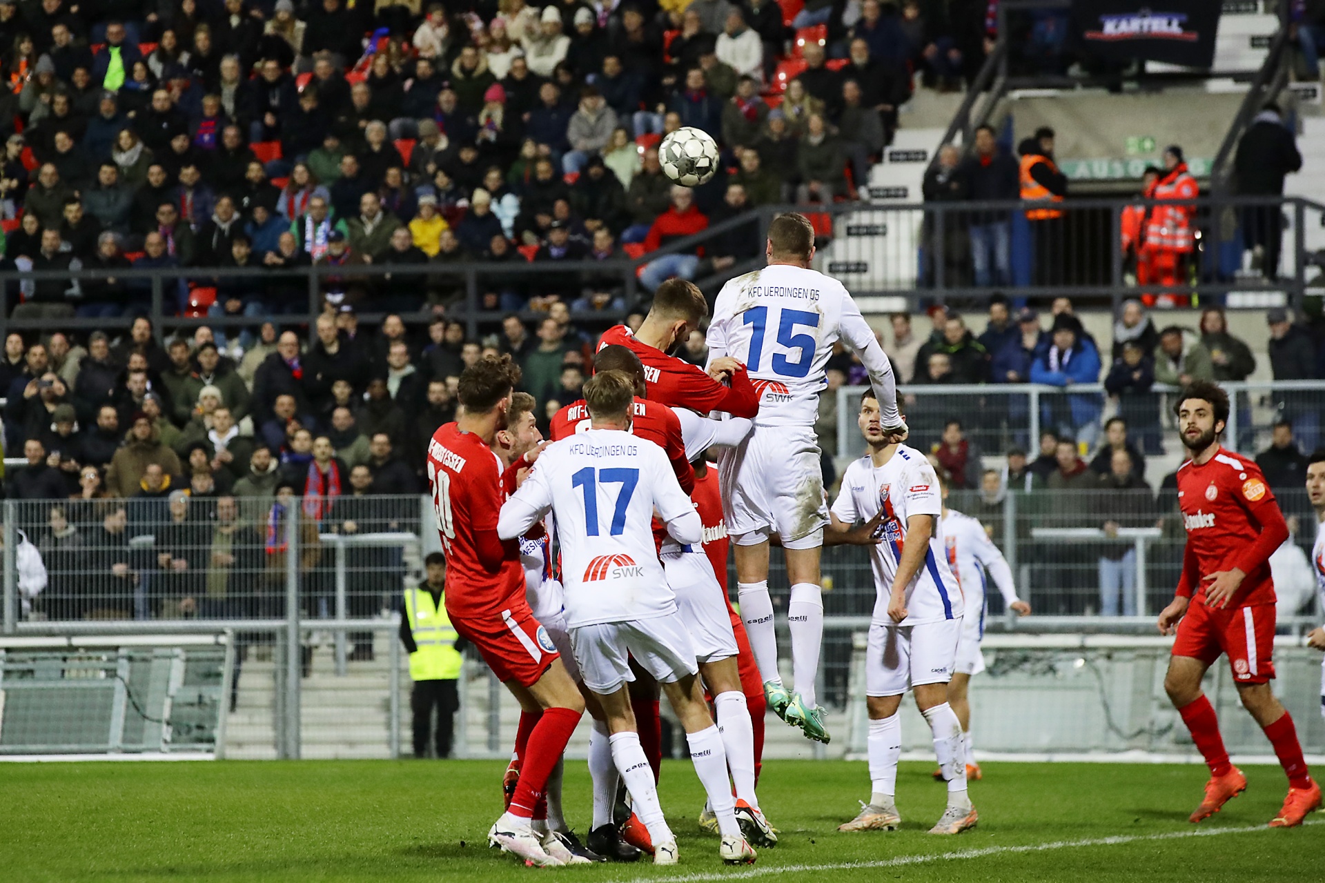[{"clang_id":1,"value":"Fußballspiel: Spieler springt zum Kopfball, umgeben von Gegenspielern im Strafraum. Im Hintergrund dichtes Publikum auf den Tribünen."}]