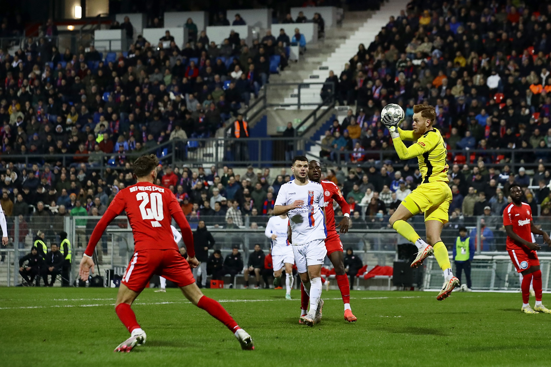 [{"clang_id":1,"value":"Fußballspiel: Torwart springt hoch, um Ball abzufangen. Spieler in Rot und Weiß im Hintergrund, Zuschauerreihen."}]