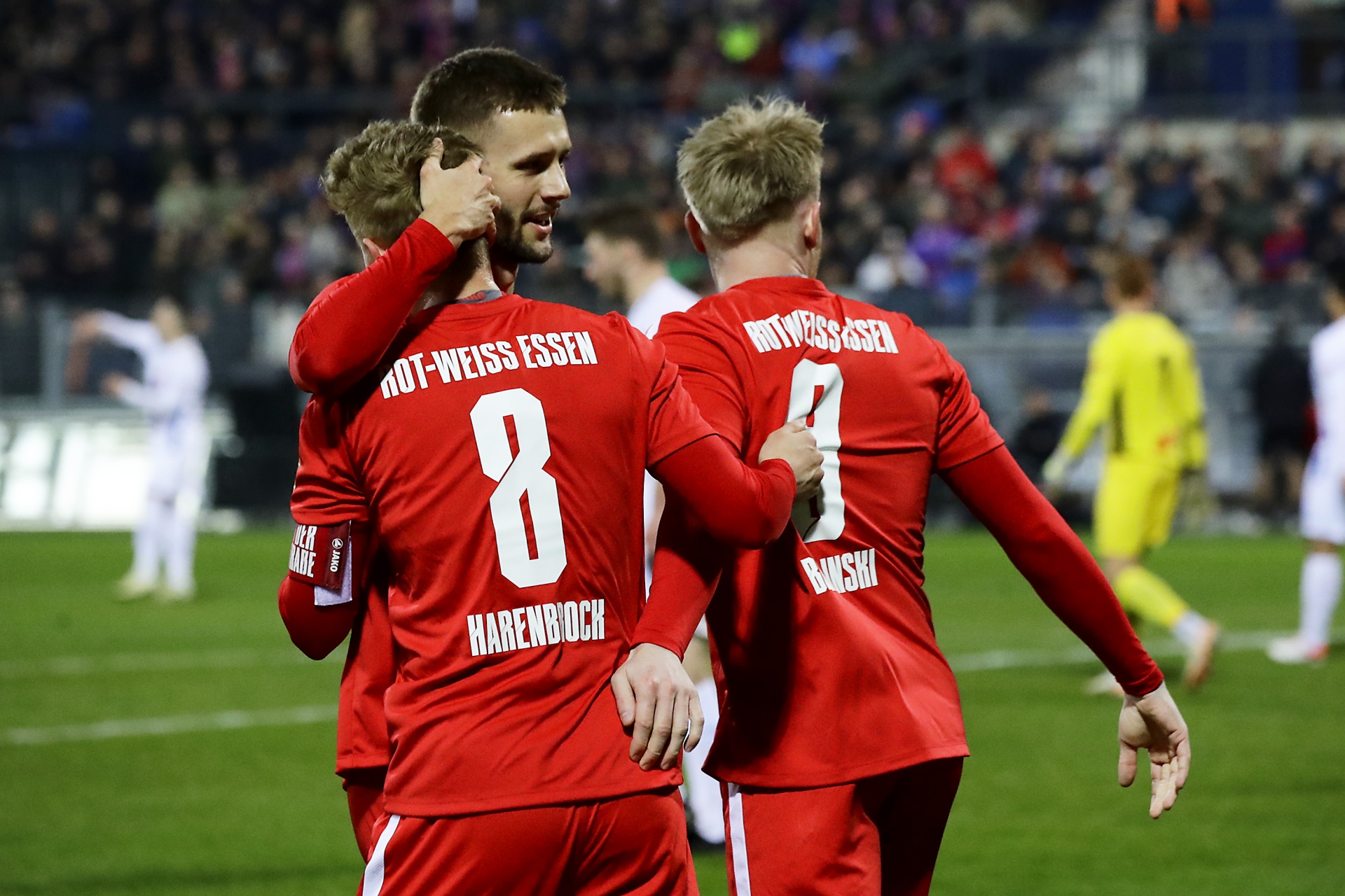 [{"clang_id":1,"value":"Rot-weiße Fußballspieler jubeln eng umschlungen auf dem Rasen. Im Hintergrund verschwommene Spieler in Gelb und die dunkle Stadionkulisse."}]