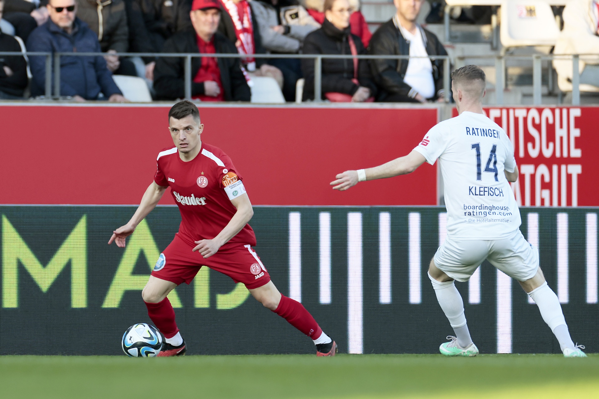 [{"clang_id":1,"value":"Zwei Fußballspieler auf grünem Rasen. Links in rot, rechts in grau-weiß. Im Hintergrund Zuschauer und Werbebanner."}]