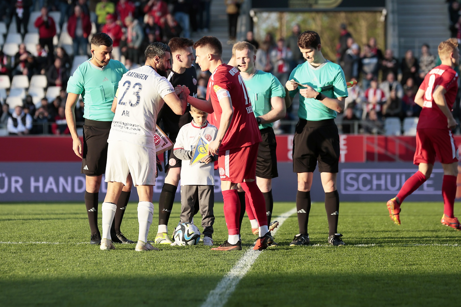 [{"clang_id":1,"value":"Fußballspiel: Spieler in rot und weiß diskutieren mit Schiedsrichtern in schwarz-weiß-gestreiften Trikots auf grünem Rasen. Zuschauerränge im Hintergrund."}]