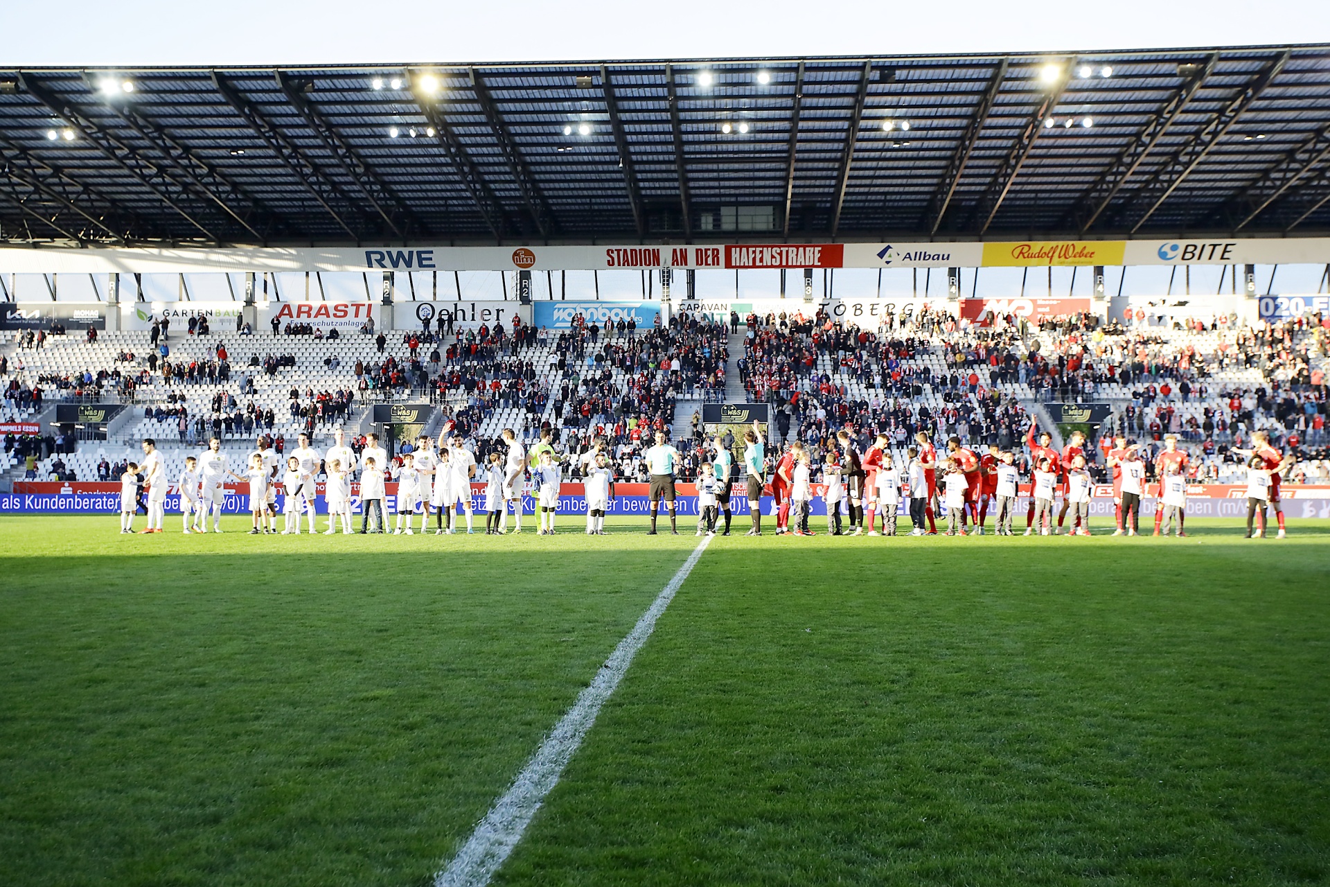 [{"clang_id":1,"value":"Fußballstadion: Spieler stehen in zwei Reihen vor vollen Zuschauerrängen. Rasenplatz im Vordergrund mit weißen Linien. Beleuchtung am Dach."}]