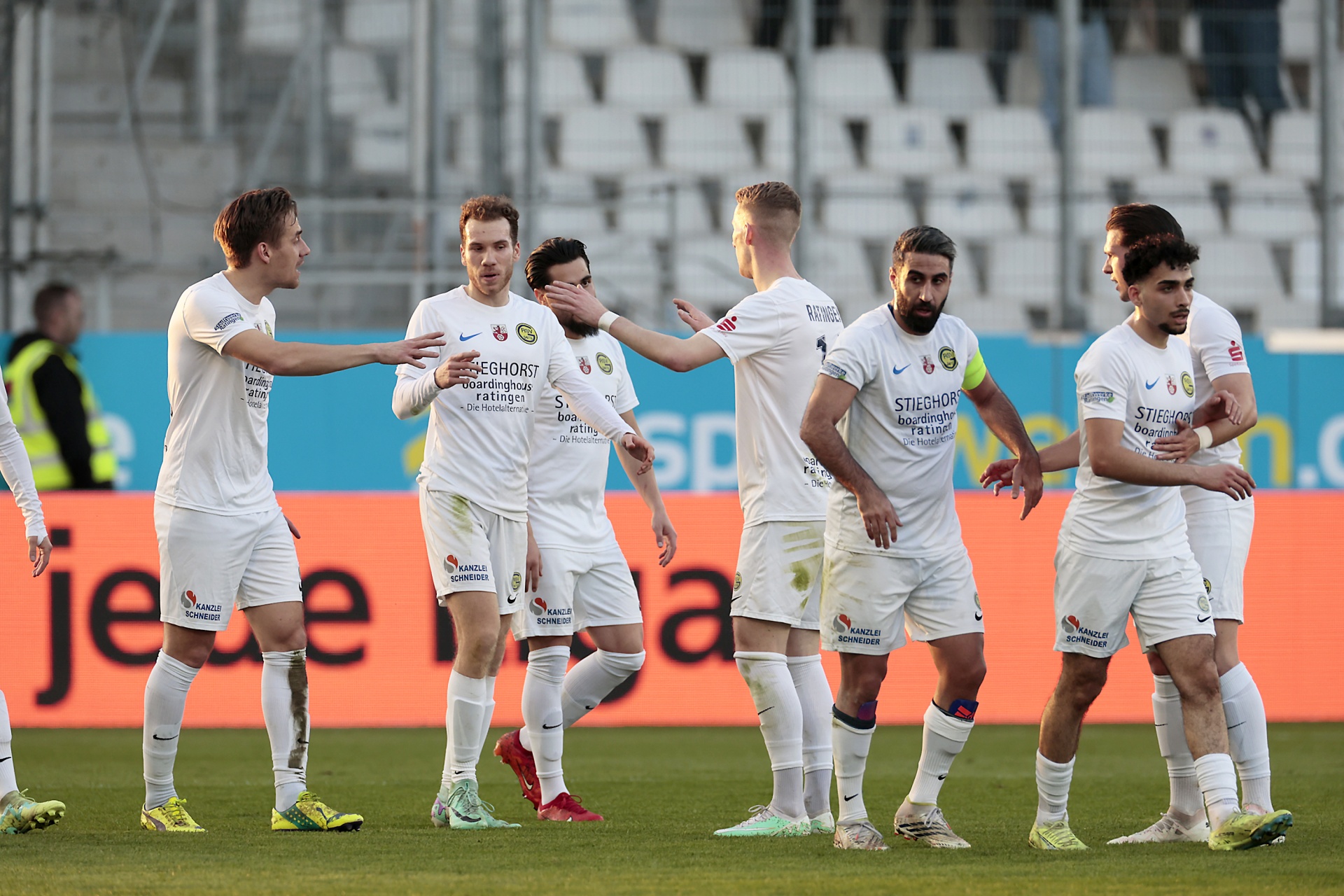 [{"clang_id":1,"value":"Fußballspieler in weißen Trikots stehen auf grünem Rasen vor einer Zuschauertribüne und jubeln gemeinsam mit erhobenen Armen."}]