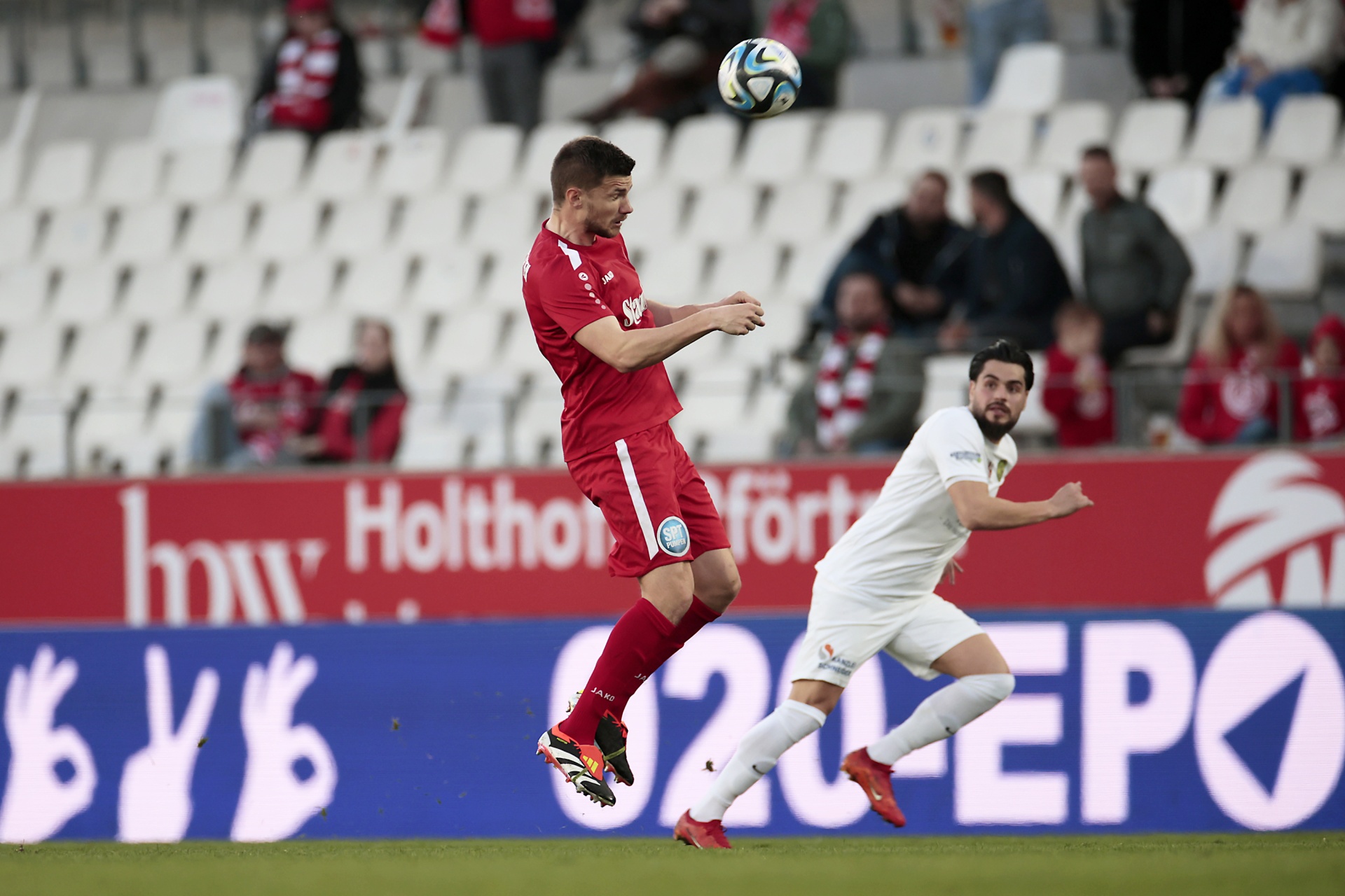 [{"clang_id":1,"value":"Zwei Fußballspieler im Spiel: Ein Athlet in Rot versucht, den Ball über einen Spieler in Weiß zu treten. Im Hintergrund Zuschauerränge."}]