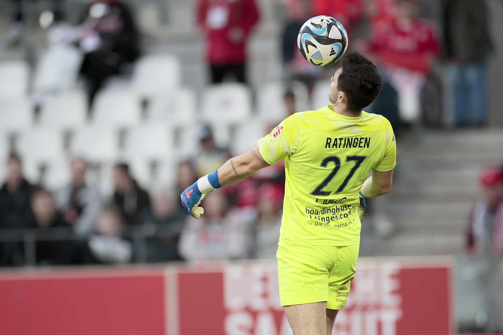 [{"clang_id":1,"value":"Handballtorwart in gelbem Trikot mit Nummer 27 balanciert Ball auf der Hand, Hintergrund Zuschauer und Werbebanner."}]