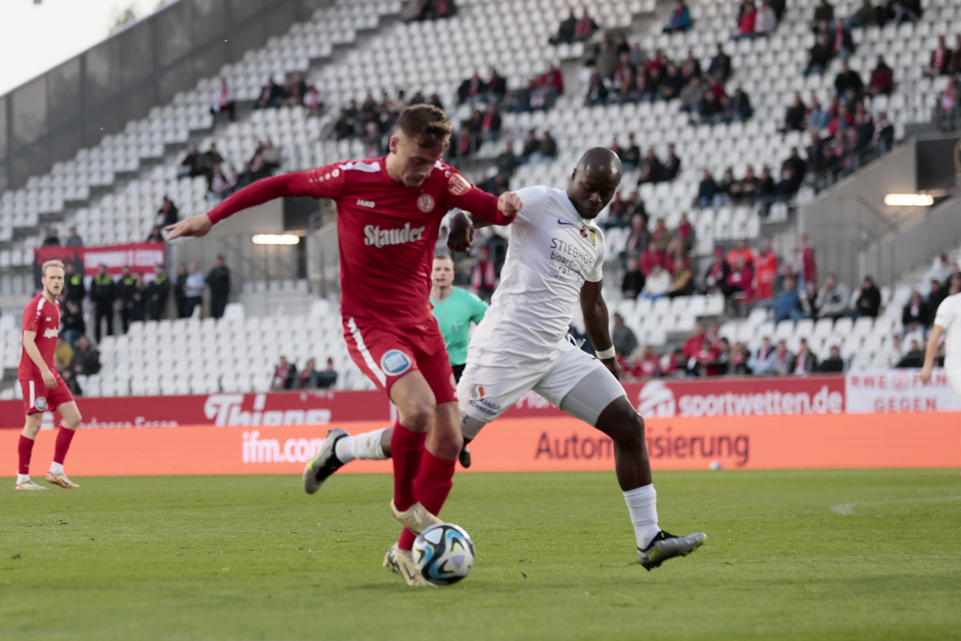[{"clang_id":1,"value":"Zwei Fußballspieler im Spiel. Links ein Spieler in roten Trikots, rechts in weißen Trikots, beide konzentriert auf den Ball. Stadion im Hintergrund."}]