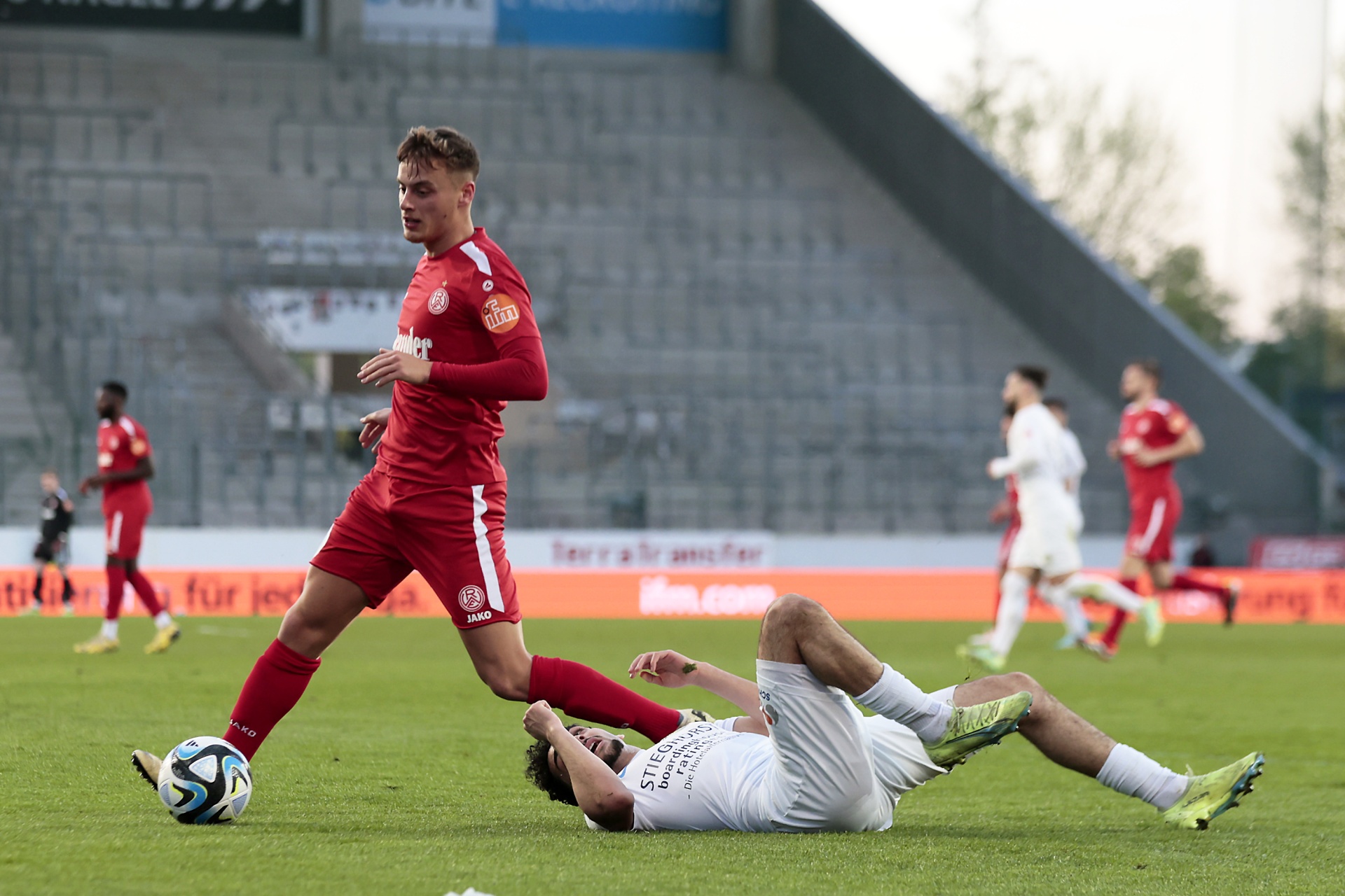 [{"clang_id":1,"value":"Fußballspiel: Spieler in roten Trikots, einer dribbelt am Ball vorbei, während ein Spieler in weißem Trikot auf dem Rasen liegt. Im Hintergrund Zuschauerreihen."}]