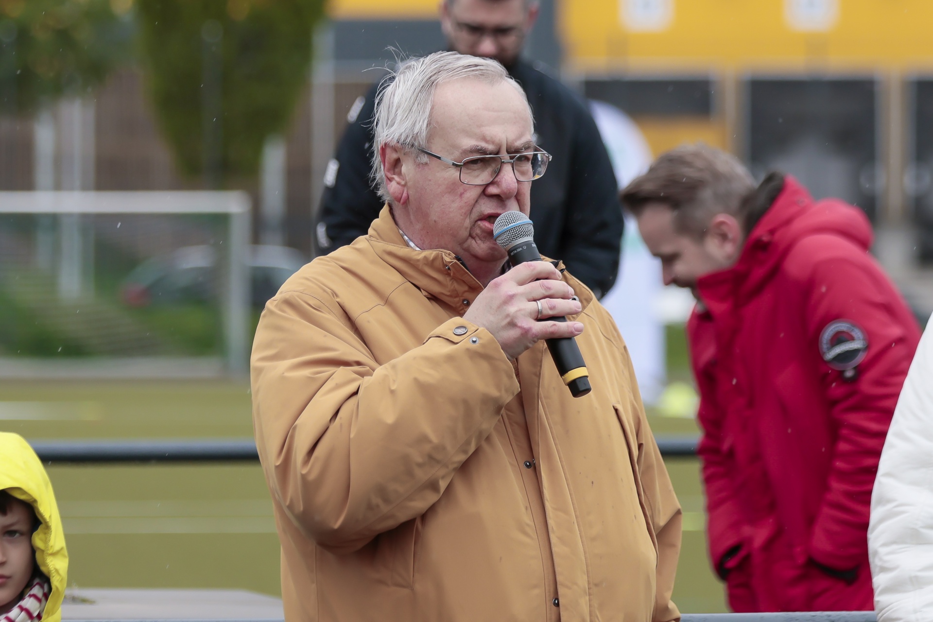 [{"clang_id":1,"value":"Älterer Mann mit Brille und beigefarbener Jacke hält Mikrofon im Freien. Im Hintergrund verschwommene Personen und ein gelber Bus."}]