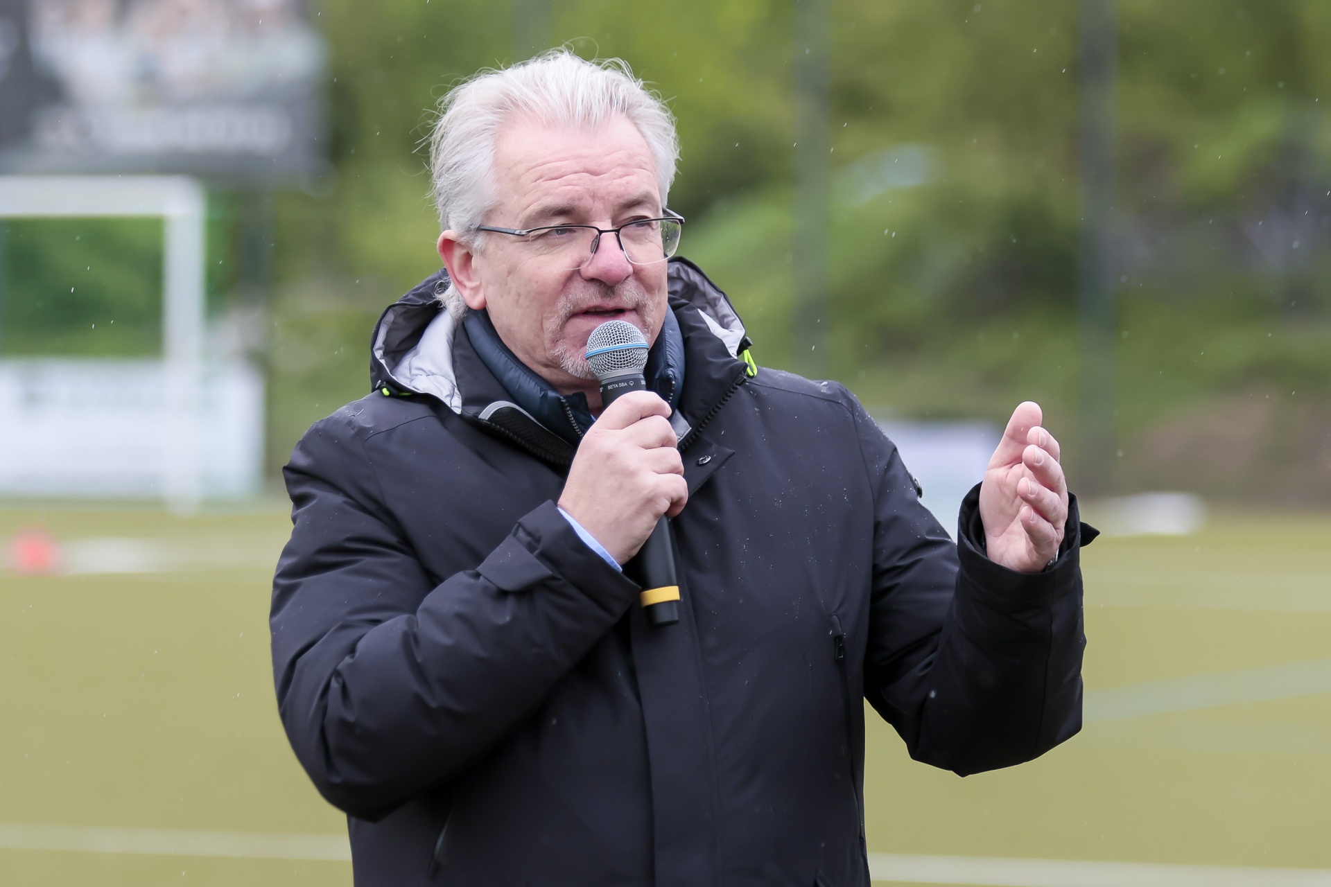 [{"clang_id":1,"value":"Mann mit grauem Haar und Brille hält Mikrofon im Freien, vermutlich während einer Rede. Im Hintergrund ein Fußballfeld und Bäume."}]