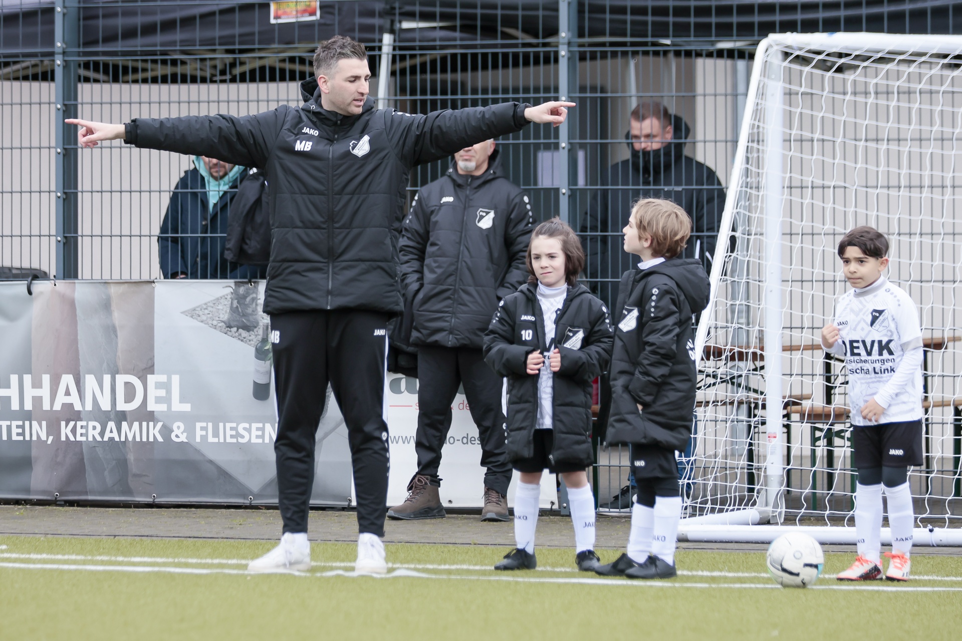 [{"clang_id":1,"value":"Fußballtrainer mit Jacke und weisenden Gesten vor Kindern in Sportkleidung am Fußballfeld. Im Hintergrund ein Zaun und Werbebanner."}]