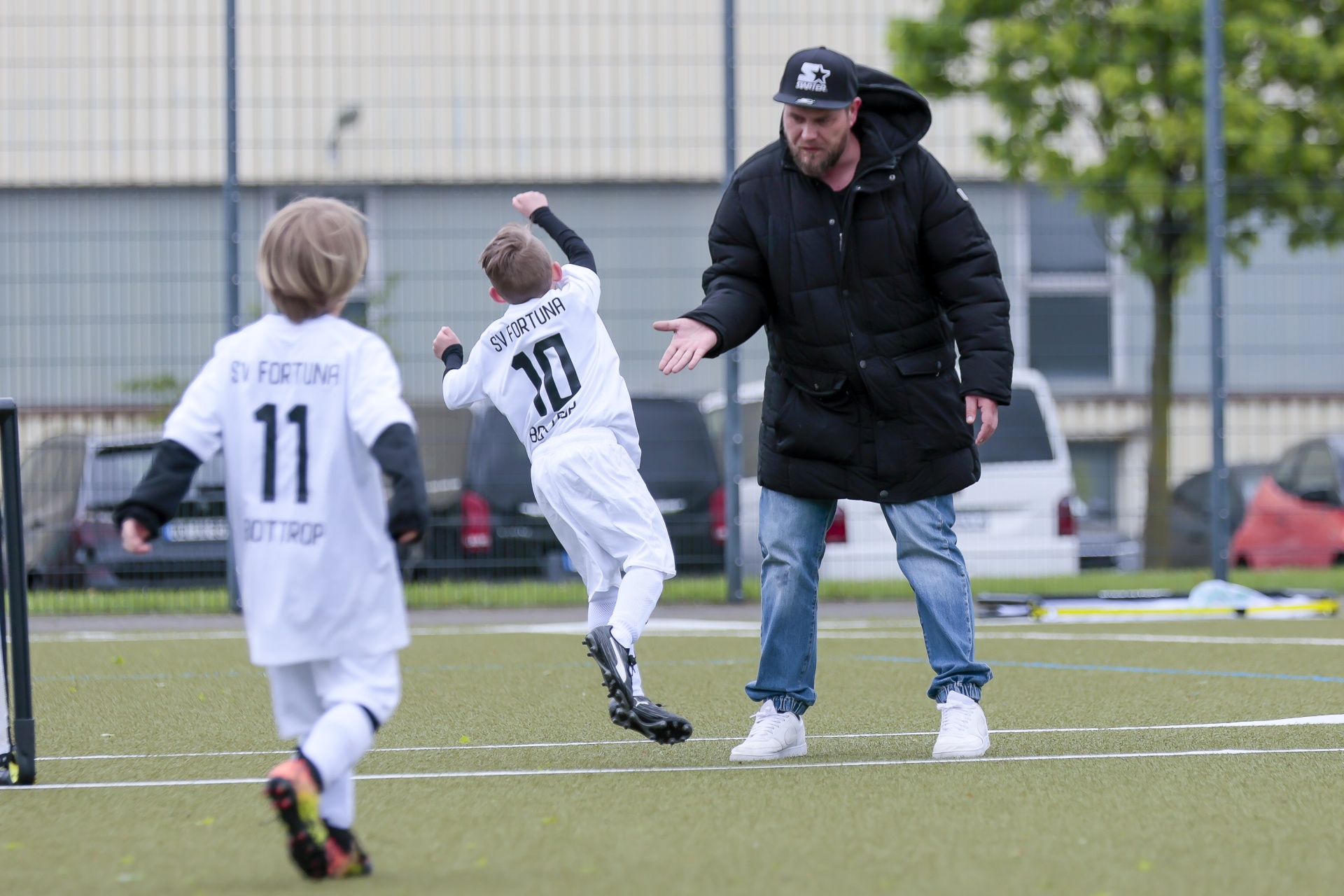 [{"clang_id":1,"value":"Zwei Fußballspieler und ein Mann auf einem Fußballfeld. Ein Kind jubelt, der Mann scheint es zu beruhigen. Im Hintergrund Gebäude und Autos."}]