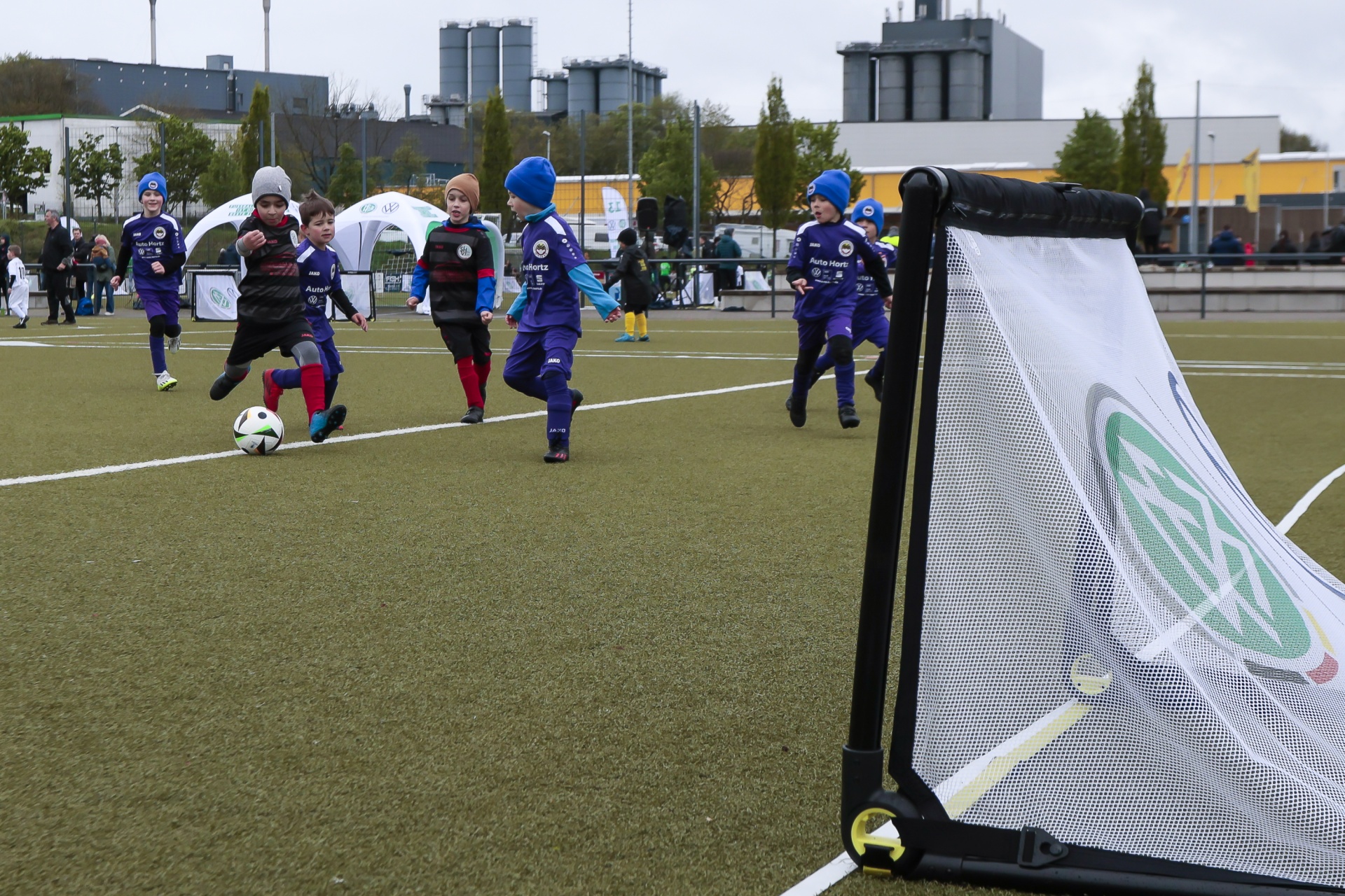[{"clang_id":1,"value":"Fußballspiel mit Kindern. Ein Spieler schießt auf das Tor, andere rennen hinterher. Im Hintergrund Gebäude und Zuschauer."}]