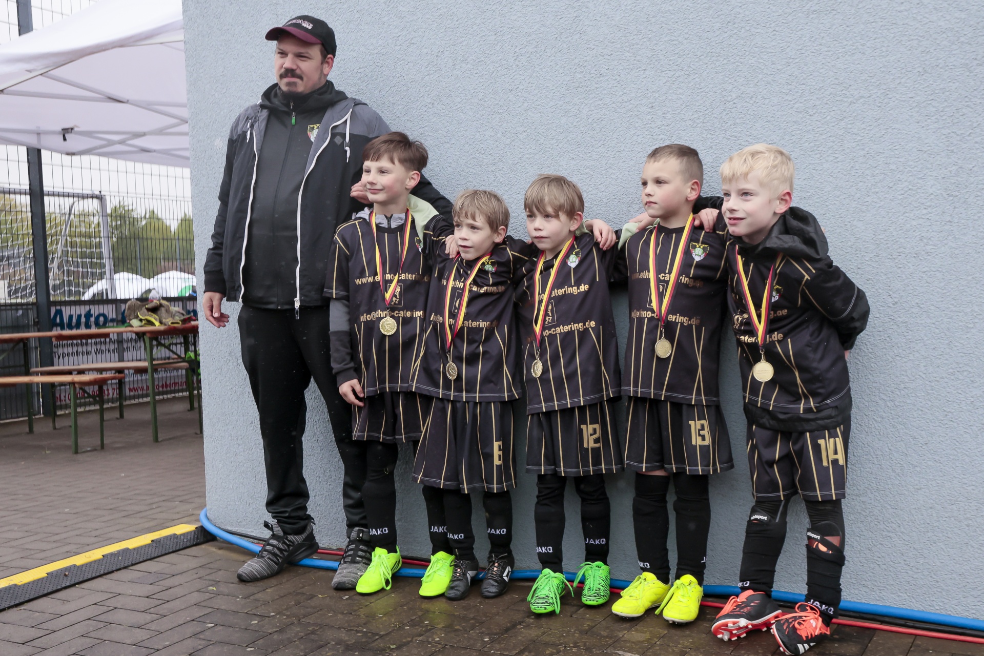 [{"clang_id":1,"value":"Fußballmannschaft mit Trainer: Sieben kleine Jungen in Fußballtrikots und Medaillen stehen mit einem Mann an einer grauen Wand."}]