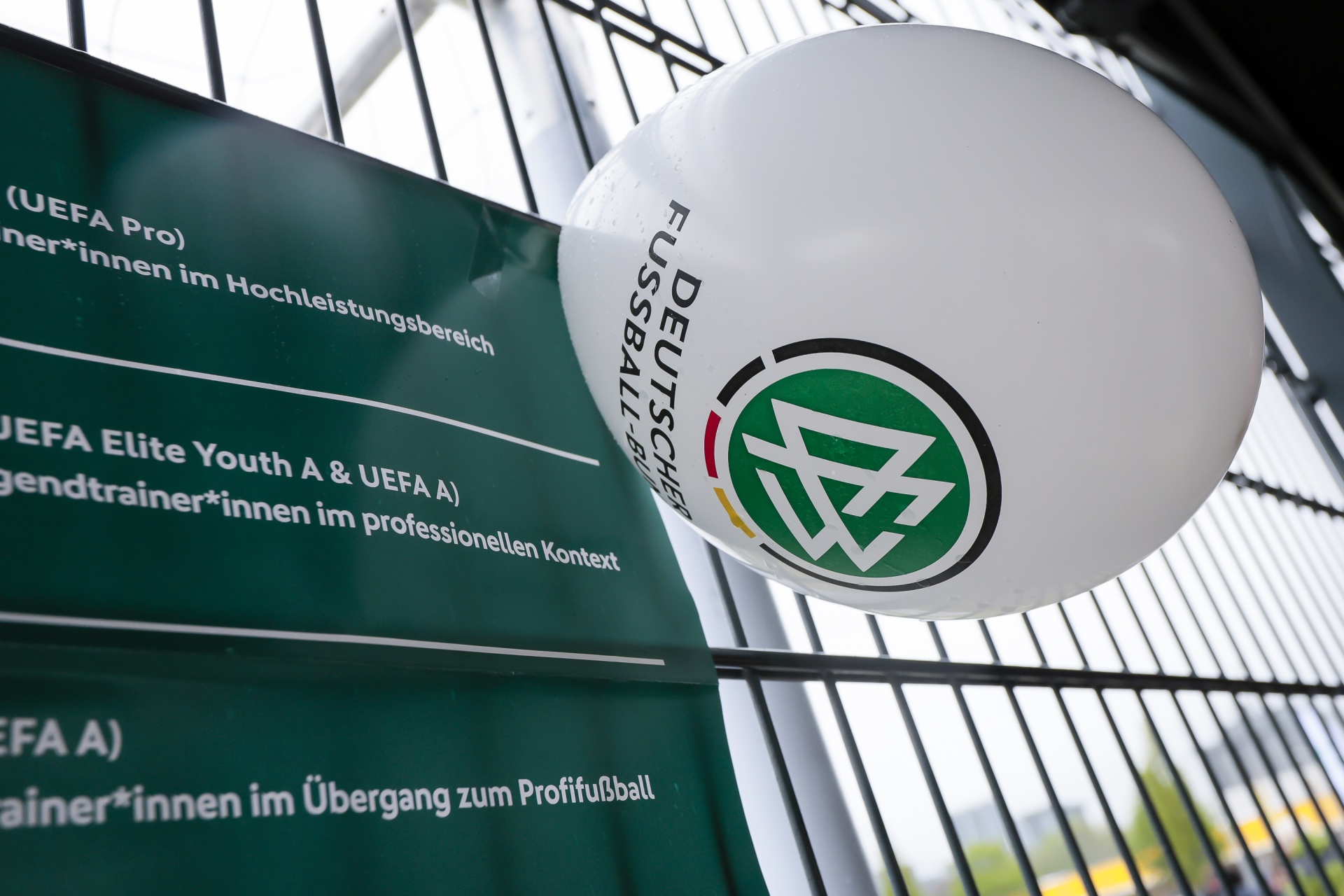 [{"clang_id":1,"value":"Fußball im Fokus: Ein weißer Fußball mit dem Logo des Deutschen Fußball-Bundes (DFB) hängt vor einer Werbetafel mit Informationen zu Trainerausbildungen."}]
