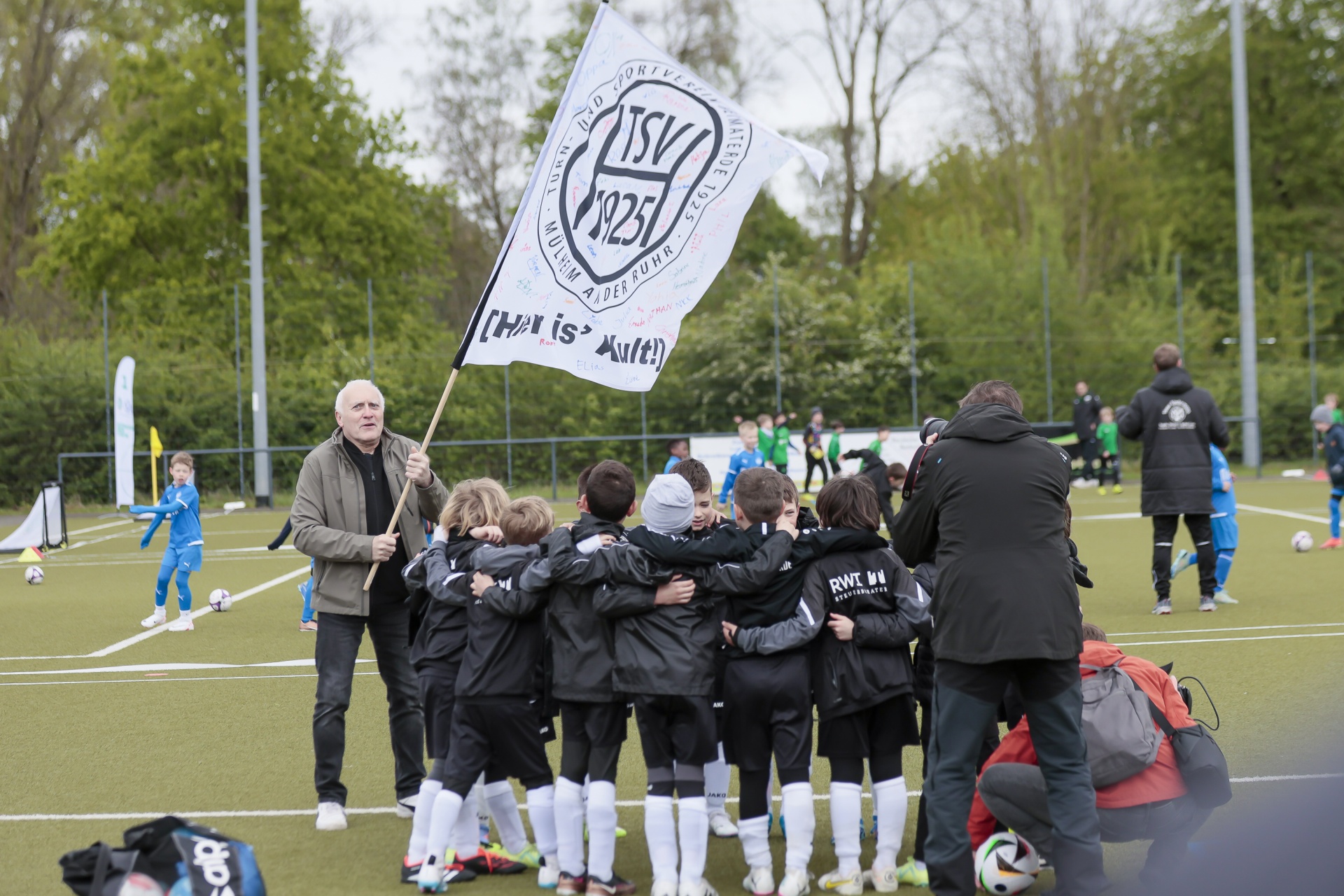 [{"clang_id":1,"value":"Fußballjugend im Kreis mit Vereinsfahne. Ein Mann reicht die Fahne, andere stehen oder sitzen im Hintergrund auf einem Fußballfeld."}]