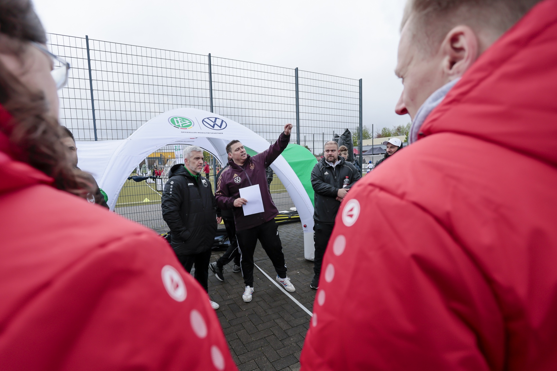 [{"clang_id":1,"value":"Fußballtrainer erklärt Taktik im Freien. Mehrere Spieler in roten Jacken stehen im Kreis um ihn, ein Torbogen mit Sponsorlogo im Hintergrund."}]