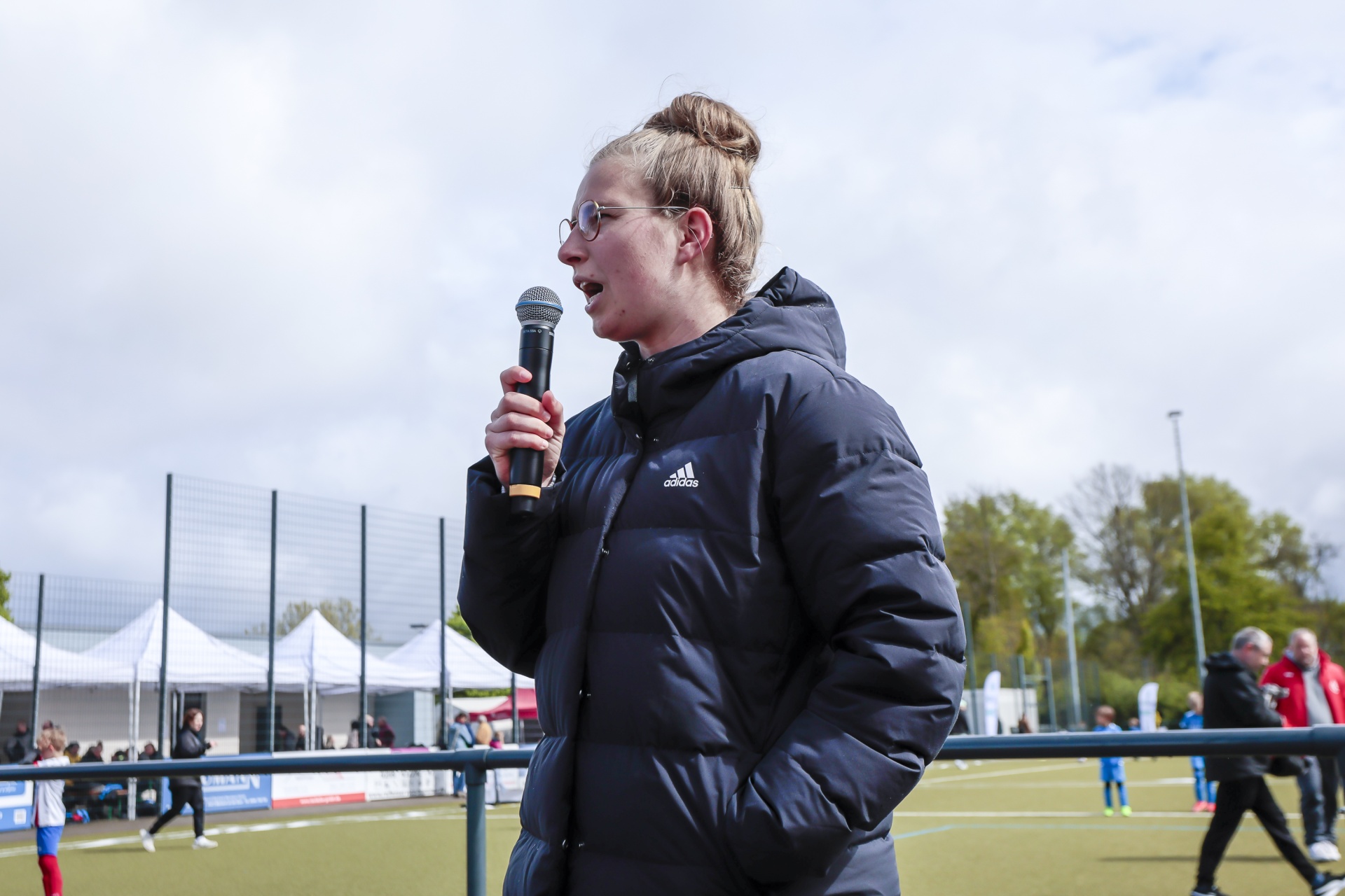 [{"clang_id":1,"value":"Frau mit hochgestecktem Haar hält Mikrofon im Freien. Sie trägt eine dunkle Jacke mit Adidas-Logo. Im Hintergrund Sportfeld und Zuschauer."}]
