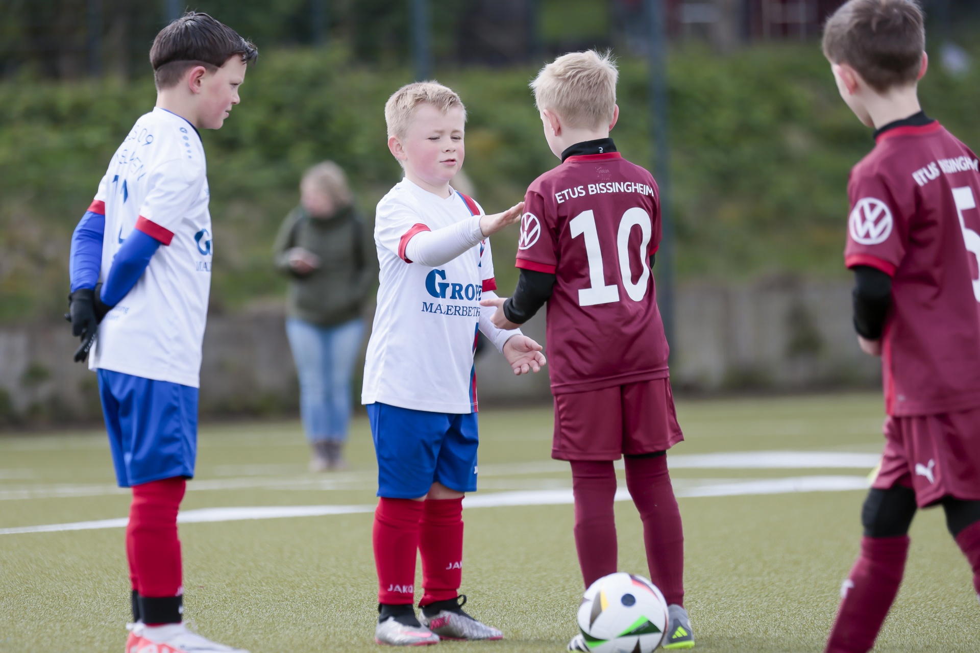 [{"clang_id":1,"value":"Vier junge Fußballspieler stehen auf einem Rasenplatz. Zwei Kinder reichen sich die Hände, während zwei weitere im Hintergrund stehen."}]