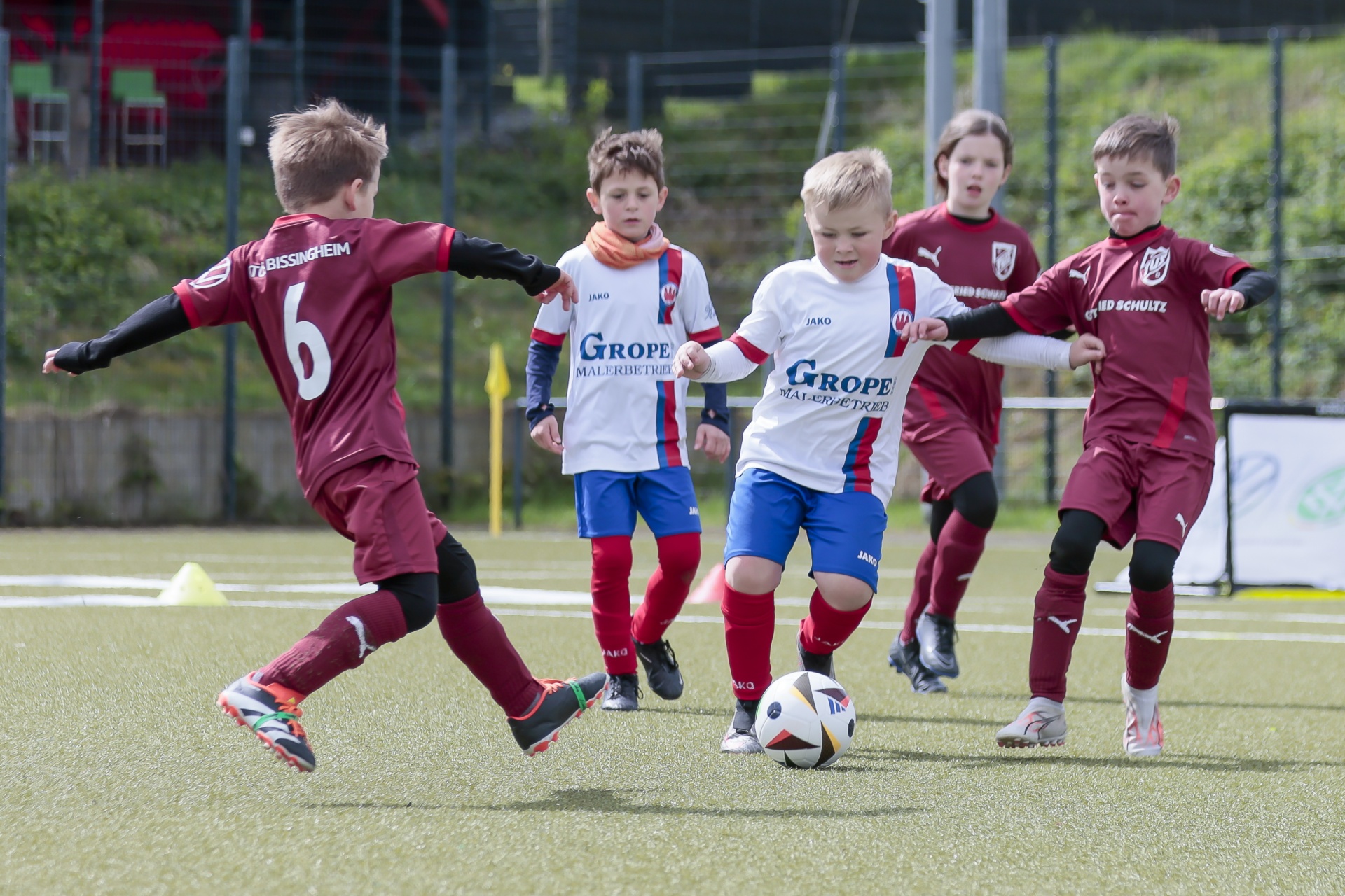 [{"clang_id":1,"value":"Jugendfußballspiel: Mehrere Kinder in Vereinskleidung kämpfen um einen Ball auf einem Rasenplatz. Rote und blau-rote Trikots dominieren. Im Hintergrund Zuschauer und ein Zaun."}]
