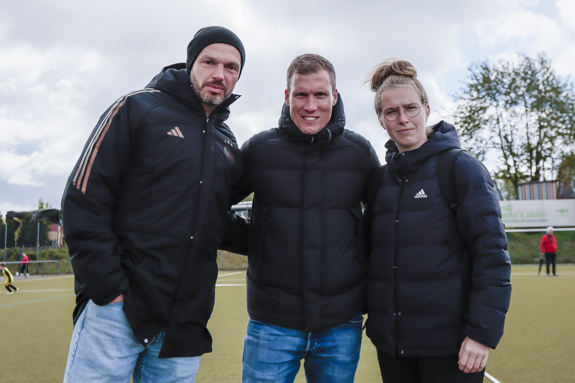 [{"clang_id":1,"value":"Drei Personen posieren vor einem Fußballfeld. Links ein Mann mit Bart und schwarzer Mütze, in der Mitte und rechts jeweils ein Mann und eine Frau, alle tragen Sportkleidung."}]