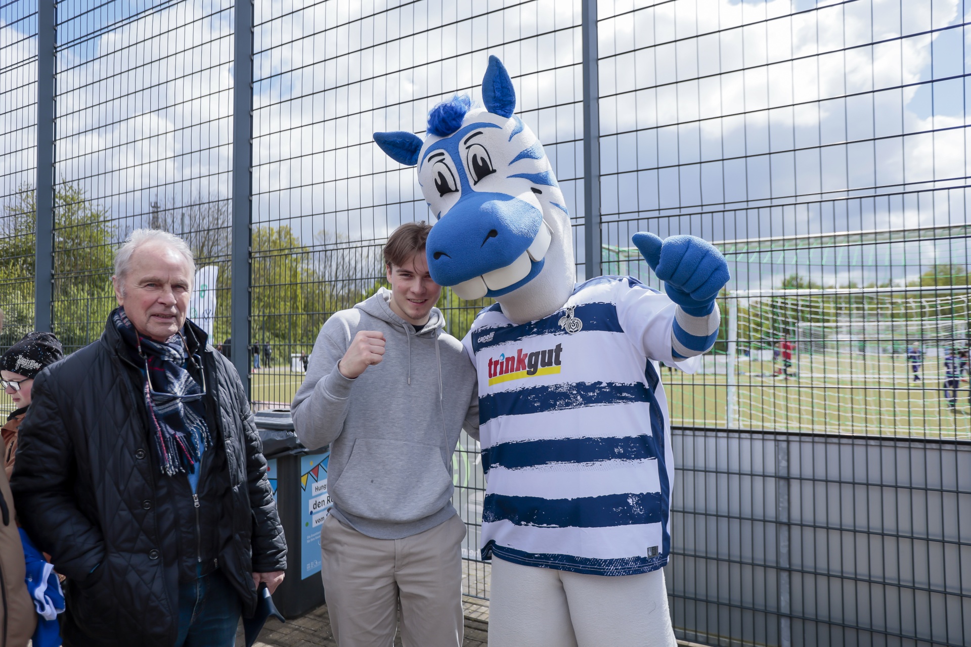 [{"clang_id":1,"value":"Zwei Männer und ein blaues Zebra-Maskottchen stehen vor einem Maschendrahtzaun. Im Hintergrund ein Fußballfeld und Zuschauer."}]
