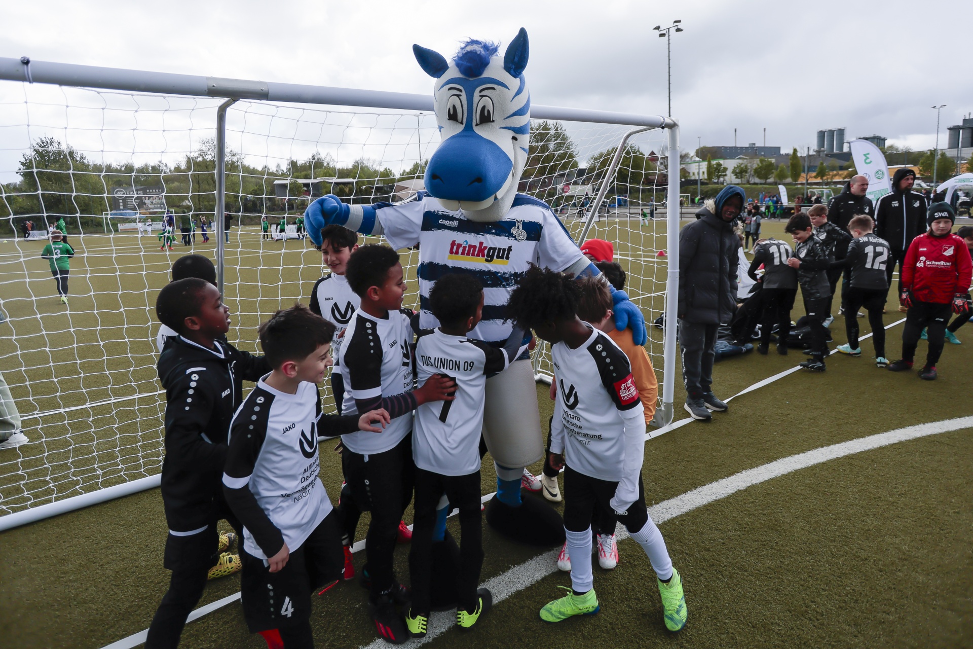 [{"clang_id":1,"value":"Fußballmannschaft mit Maskottchen. Eine Gruppe junger Fußballspieler umarmt ein blaues Pferdemaskottchen vor einem Fußballtor auf einem Kunstrasenplatz."}]